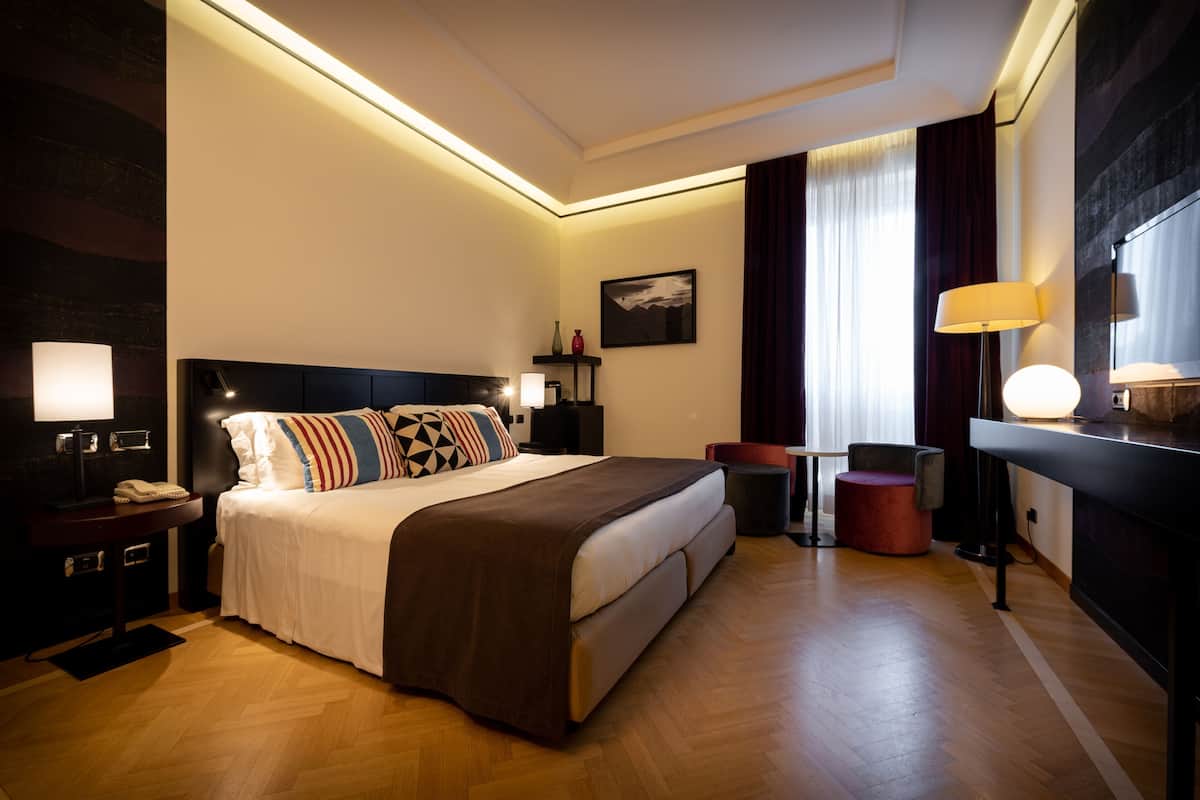 A hotel room with a bed, bedside table, lamp, chair, and a desk with a lamp.