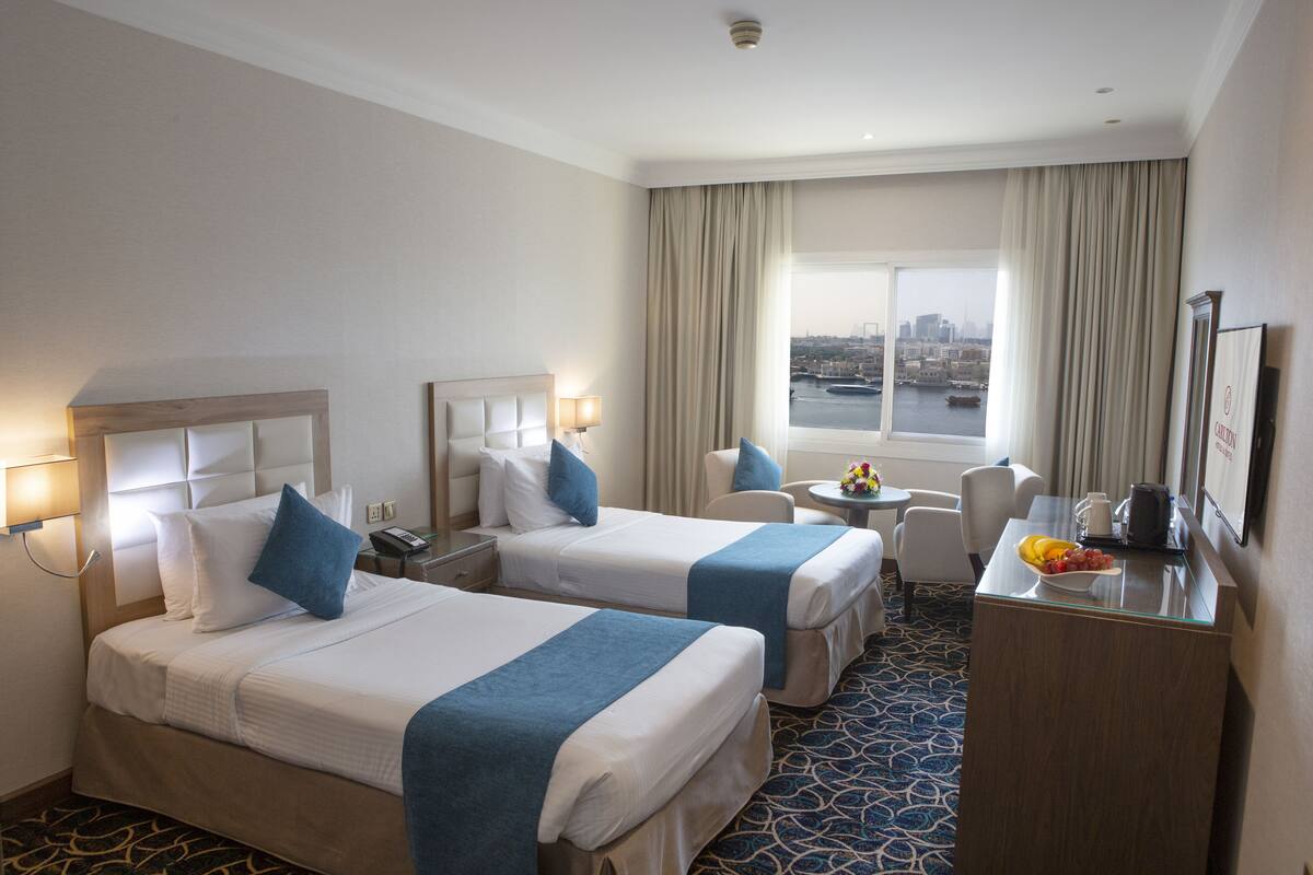 A hotel room with two beds, a desk, a chair, a small table, and a view of the city through large windows.