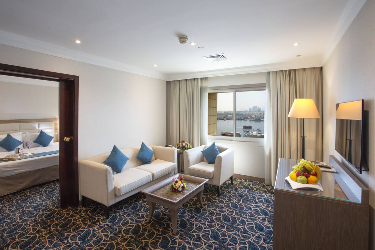 A hotel room with a bed, two sofas, a coffee table, a desk with a lamp, and a large window with a view of the city.