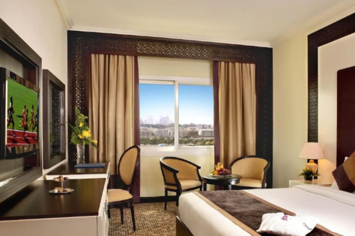 A hotel room with a bed, two chairs, a desk, a TV, and a window with a city view.