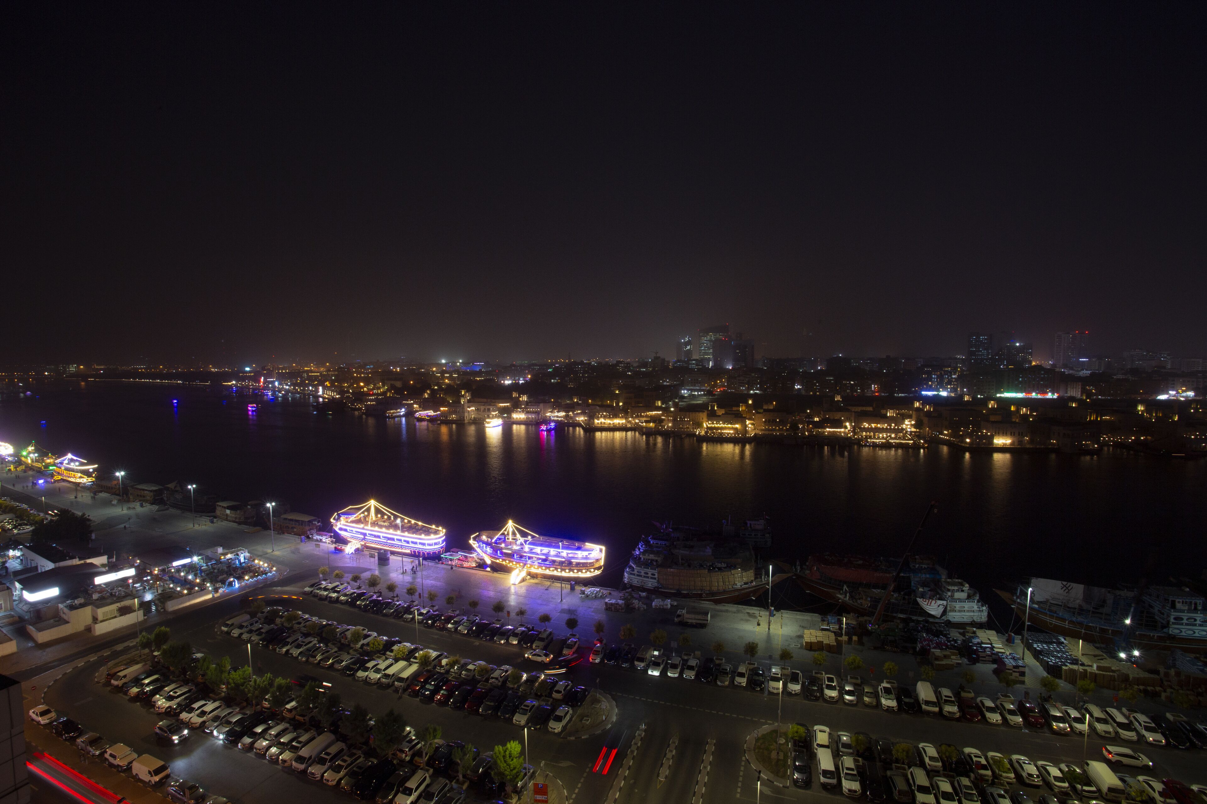 A nighttime cityscape with a waterfront area, illuminated buildings, and a parking lot.