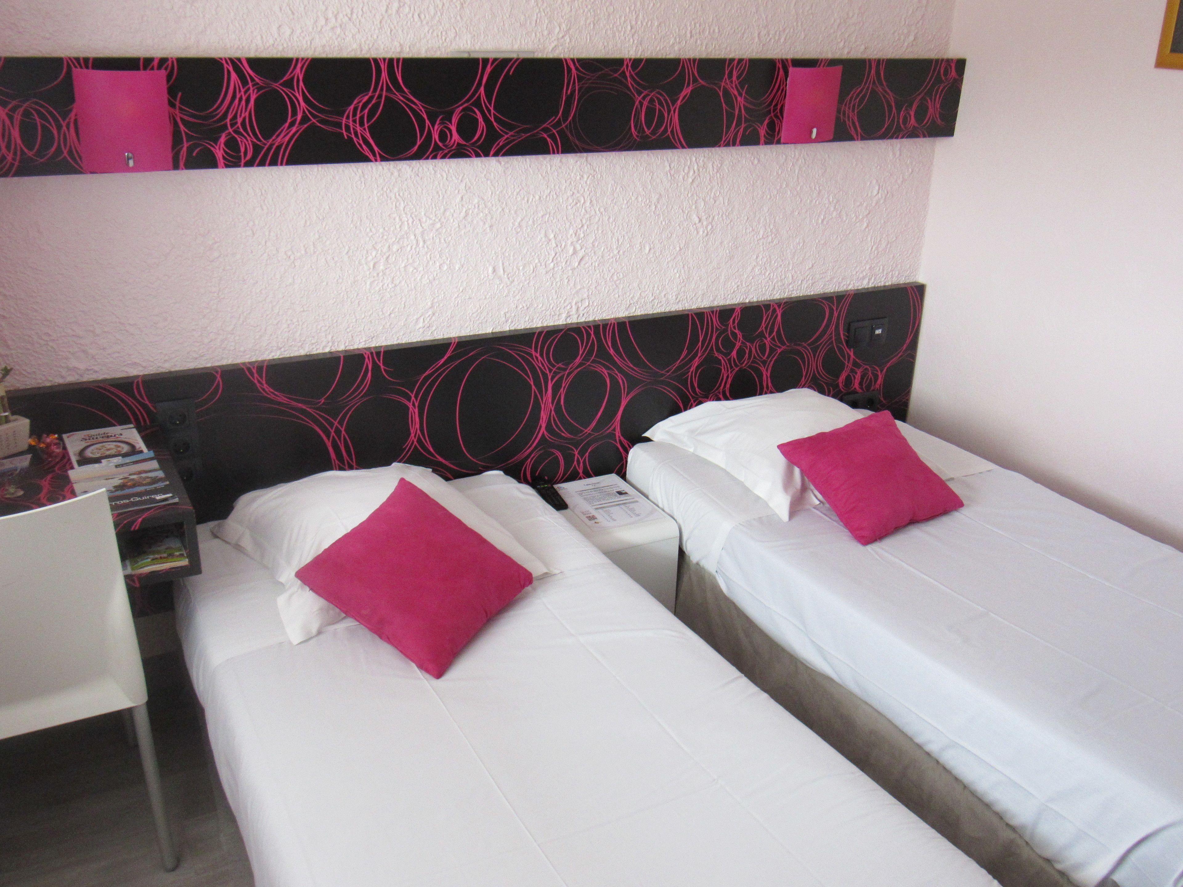Habitación de hotel con dos camas individuales, cada una con colchón blanco y almohada rosa. El cabecero presenta un diseño abstracto en negro y rosa. Hay una mesita de noche con una lámpara y algunos objetos encima.
