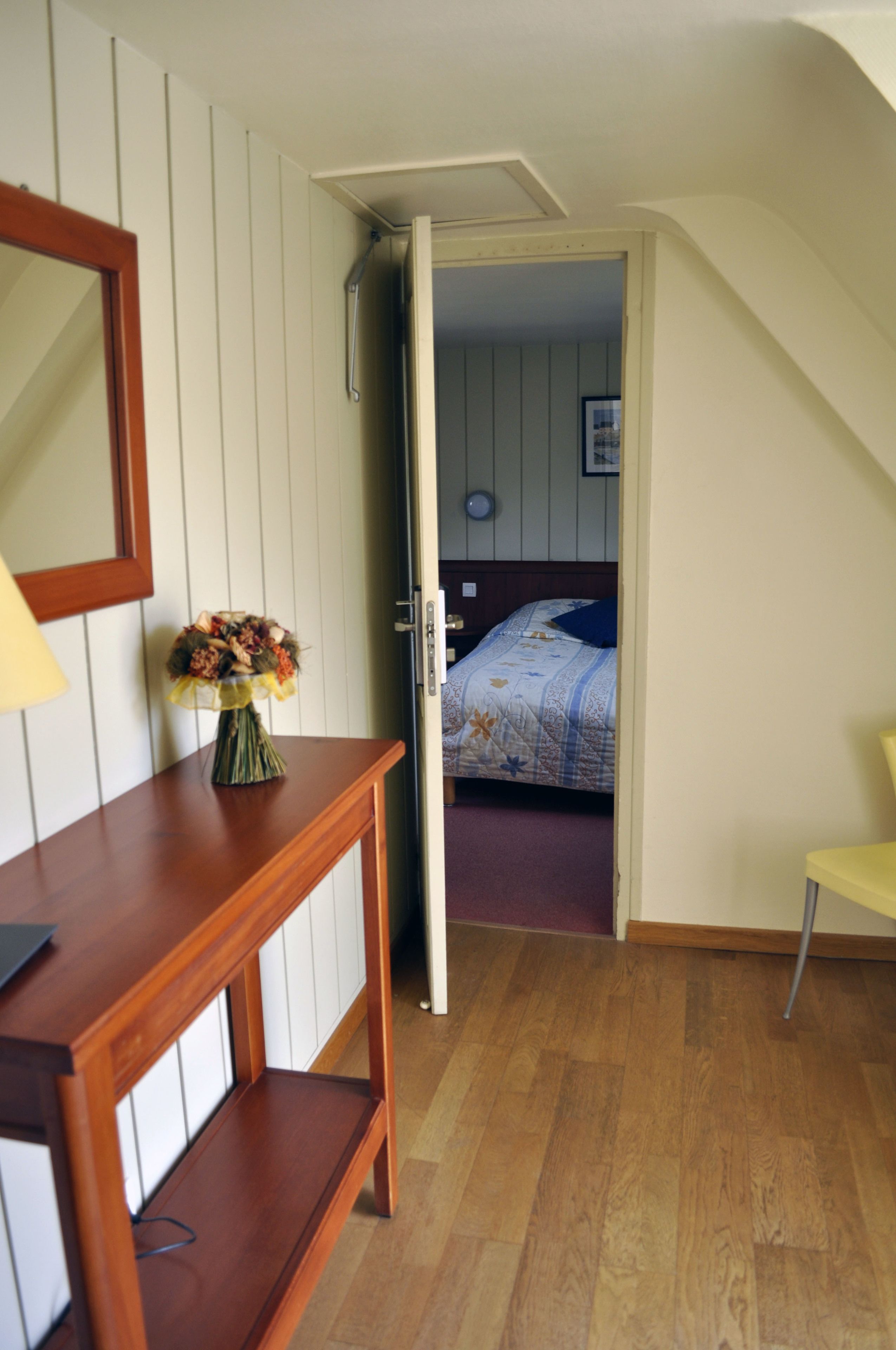 A room with a wooden desk, a vase with flowers, a bed with a blue pillow, and a chair.