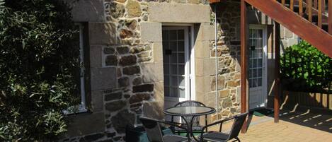 A stone building with a wooden staircase, outdoor seating with chairs and a table, and a window with a door.