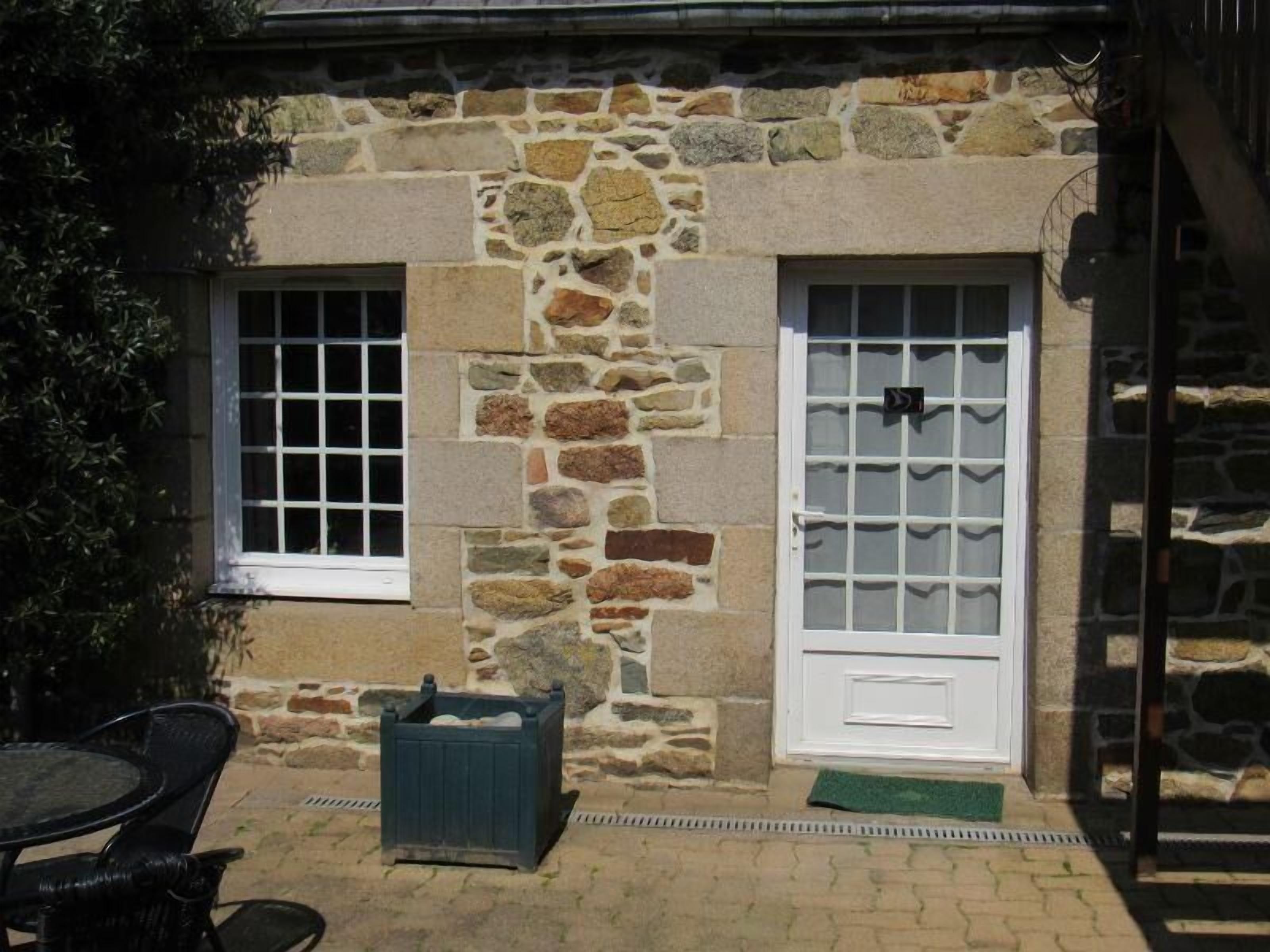 Un bâtiment en pierre avec deux fenêtres et une porte, un petit espace extérieur aménagé avec des sièges et un tapis vert à l’entrée.