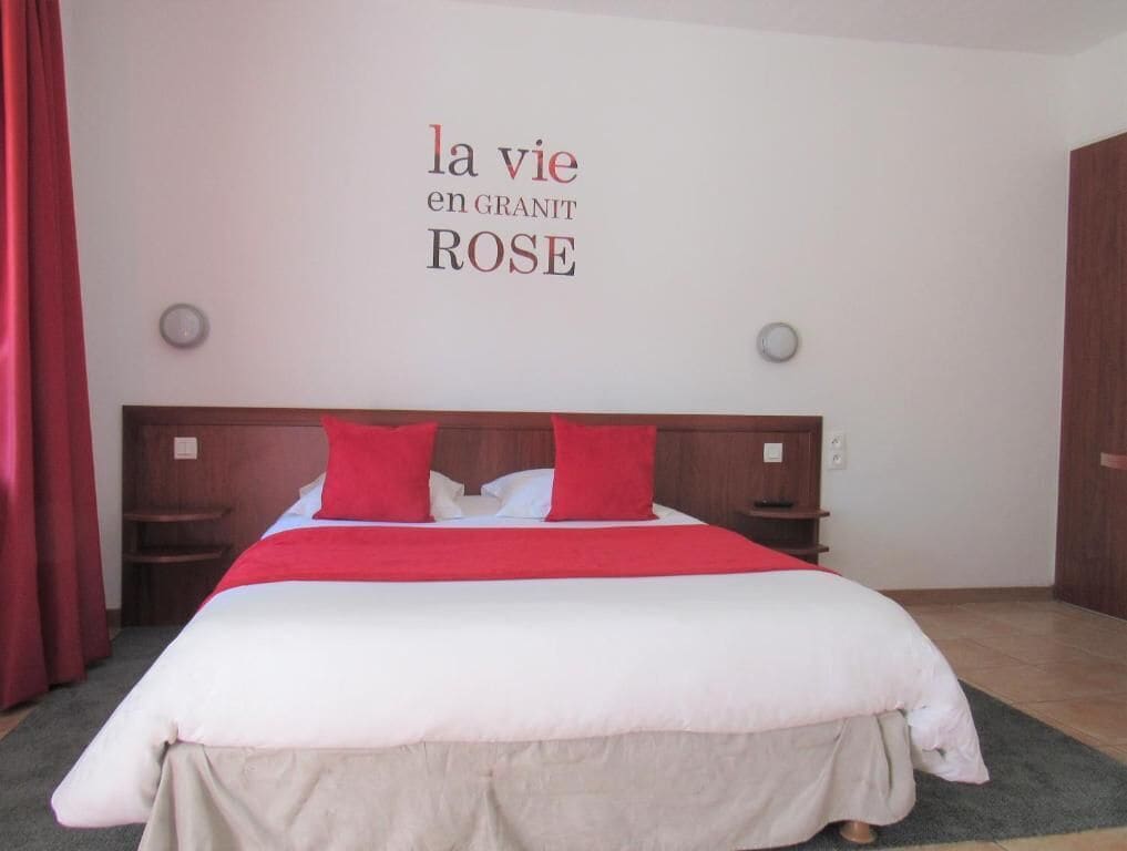 Una habitación de hotel con cama, ropa de cama roja y blanca, cabecero de madera y una pared con texto.
