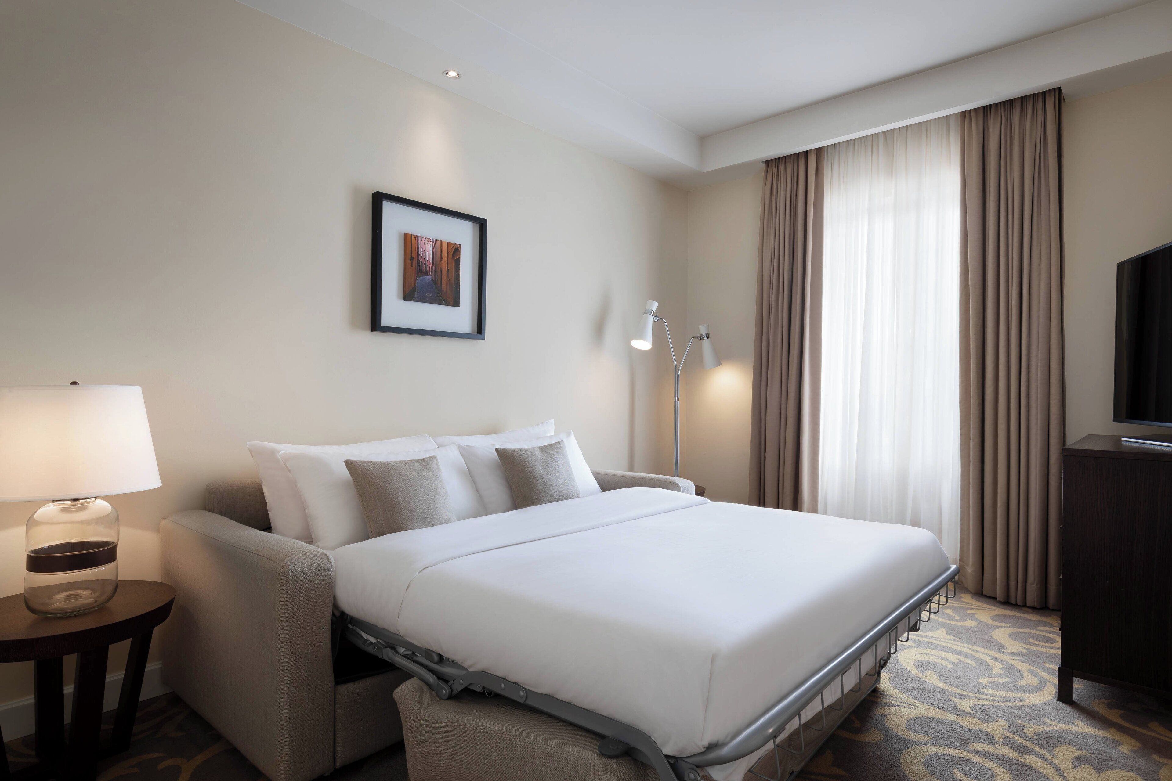 Ein Hotelzimmer mit Bett, Schreibtisch, Lampe, Fernseher und einem Bild an der Wand.