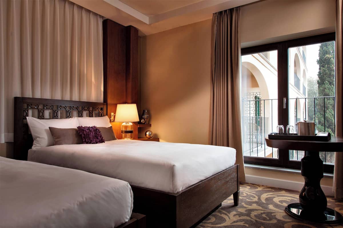 A hotel room with two beds, a nightstand with a lamp, a table with a vase, and a balcony with a view of buildings.