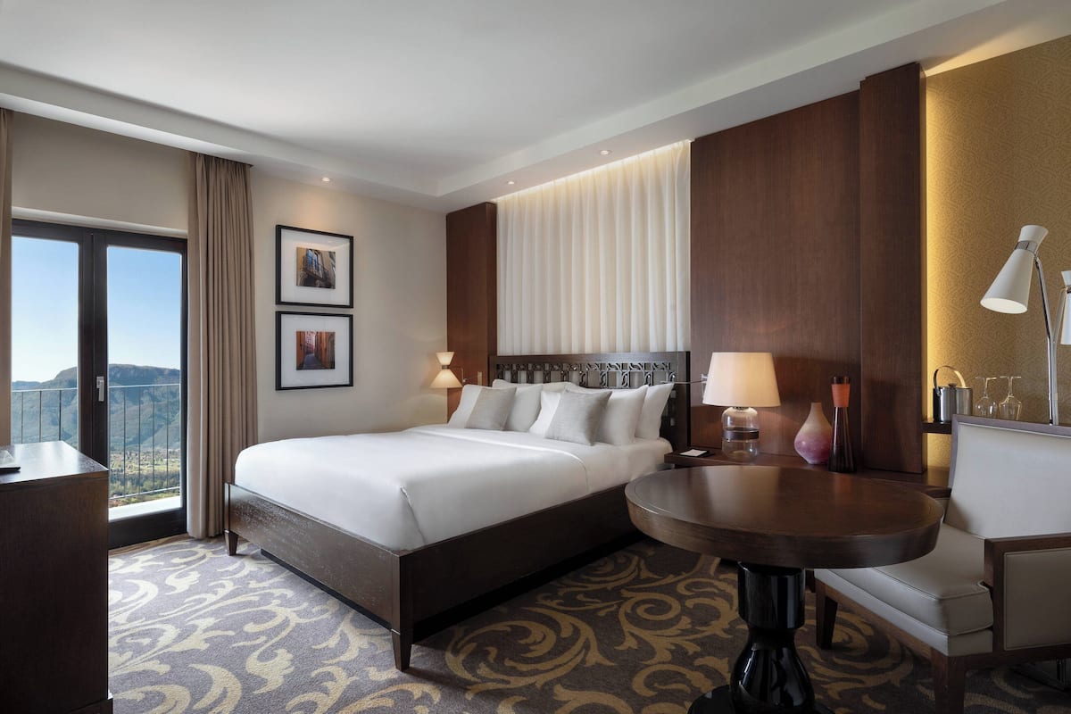 A hotel room with a large bed, a desk, a round table, and a view of the mountains.
