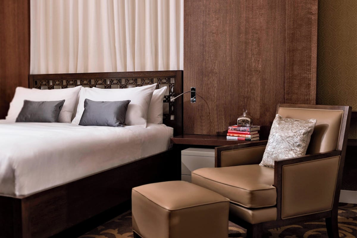 A hotel room with a large bed, a wooden headboard, a bedside table with books, a chair, and a small table with a decorative item.