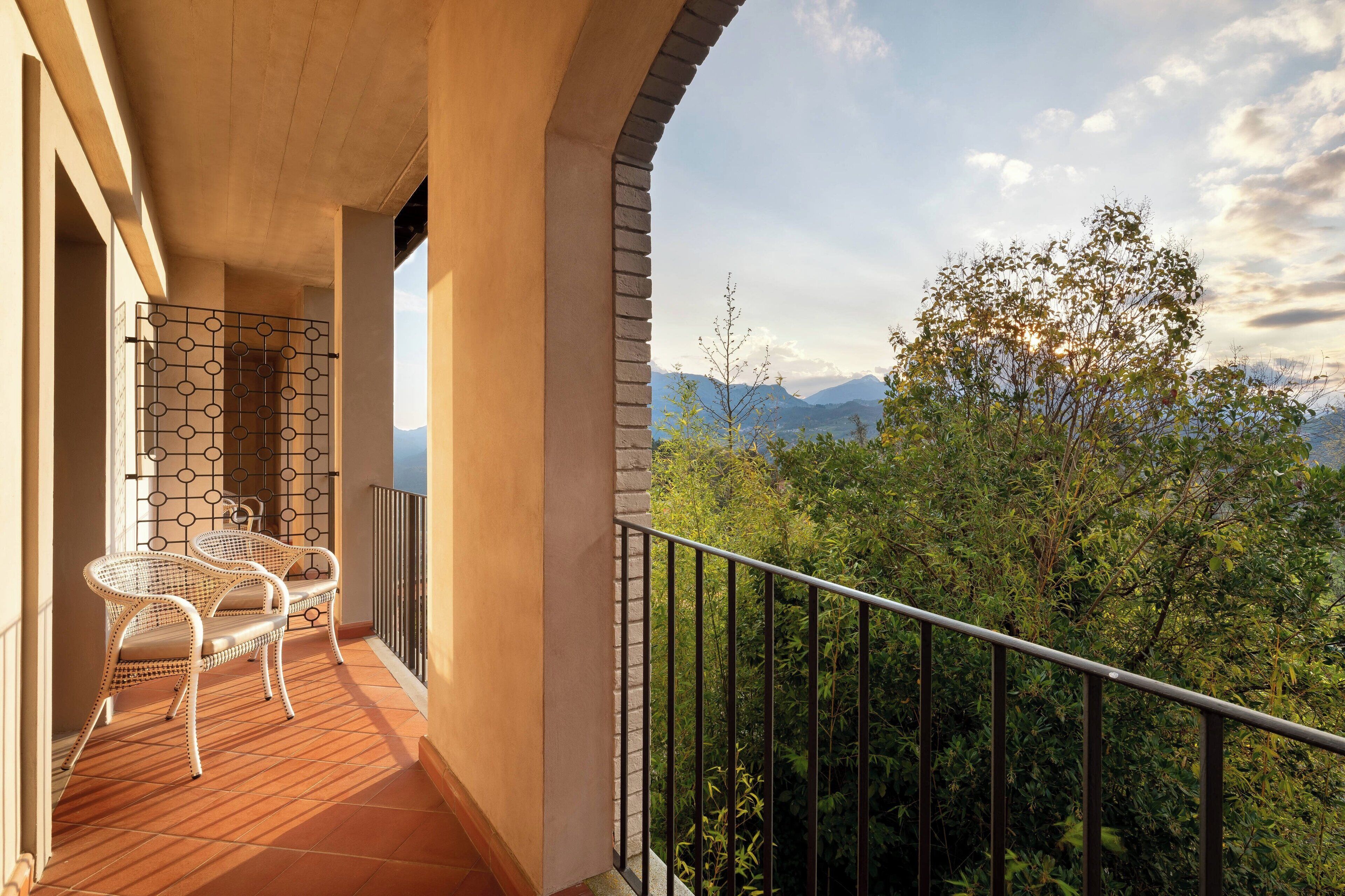 Varanda com duas cadeiras de vime, piso de azulejos e vista para árvores e montanhas.