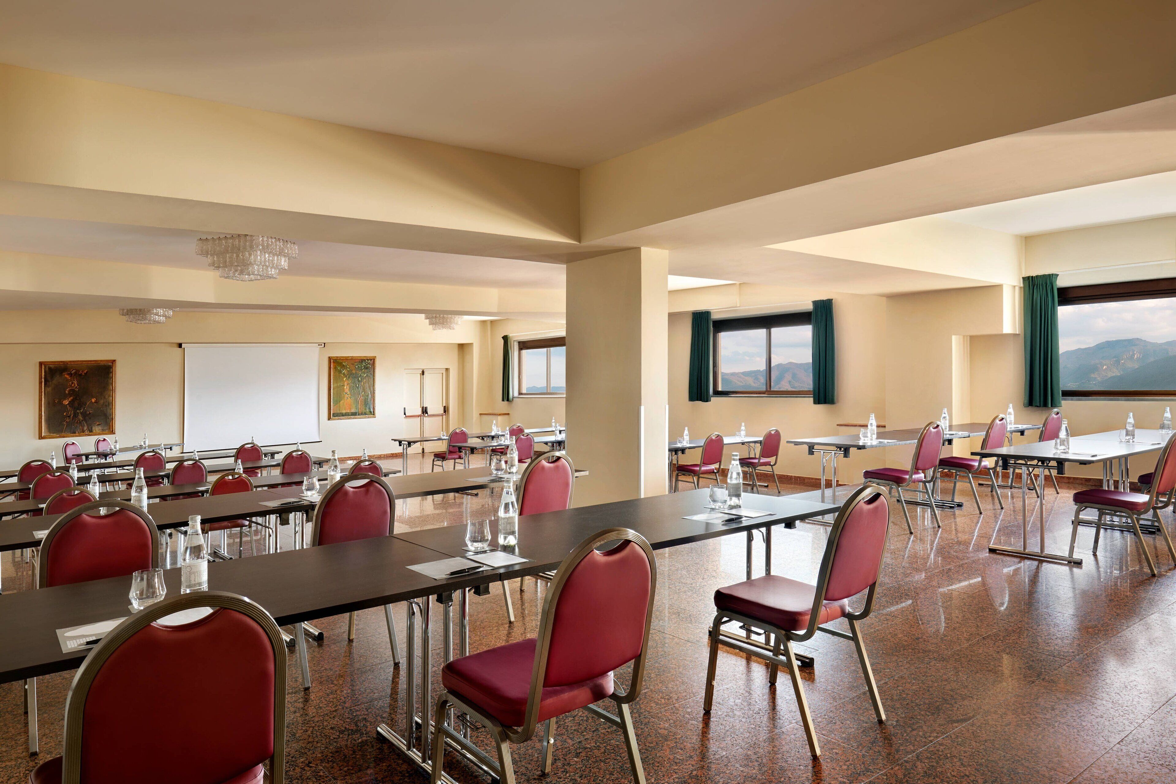 A conference room with multiple tables and chairs, large windows with curtains, and a chandelier.