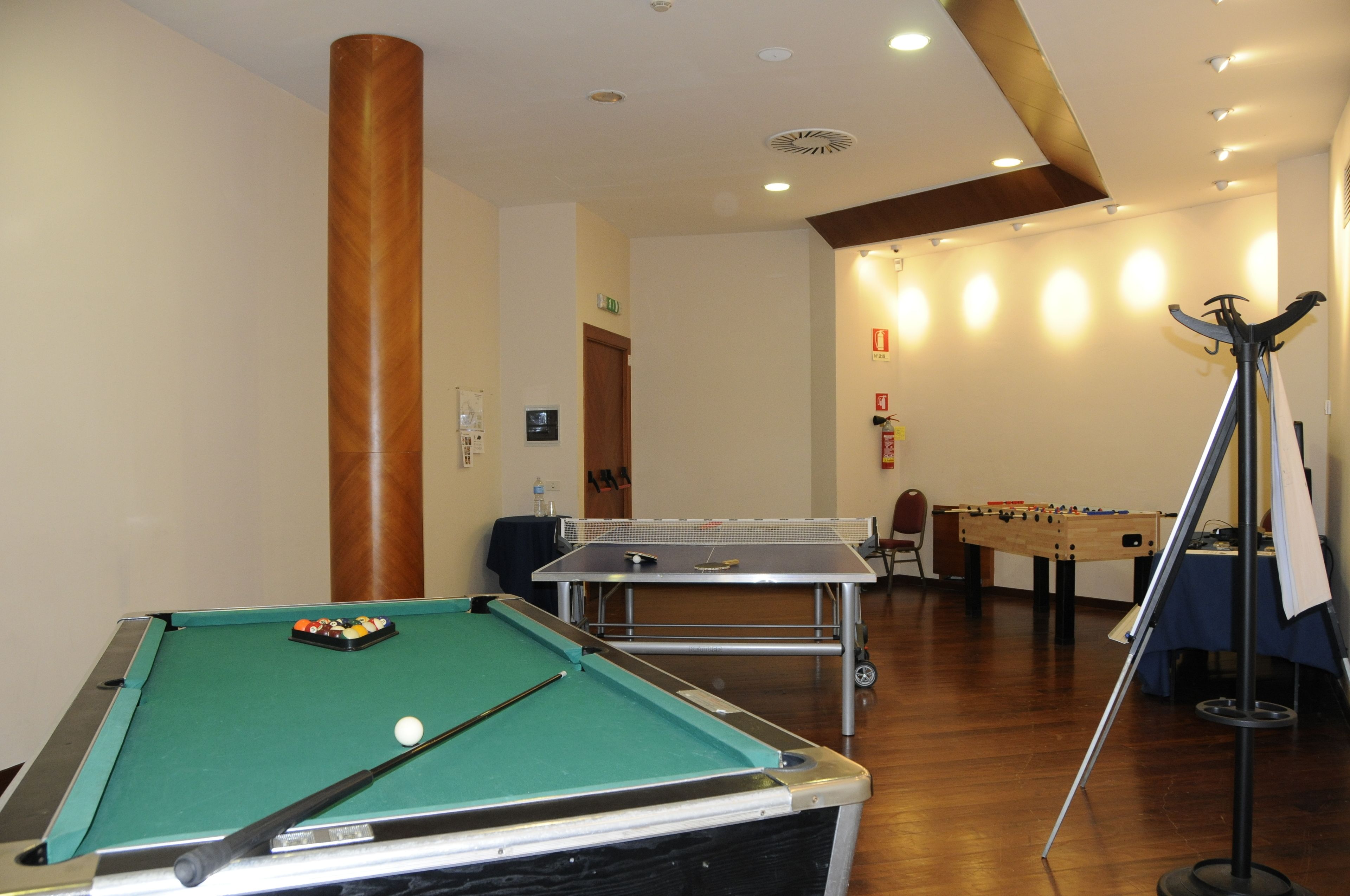 Uma mesa de bilhar, uma mesa de futebol de mesa e uma mesa de sinuca em um espaço com piso de madeira e iluminação embutida.