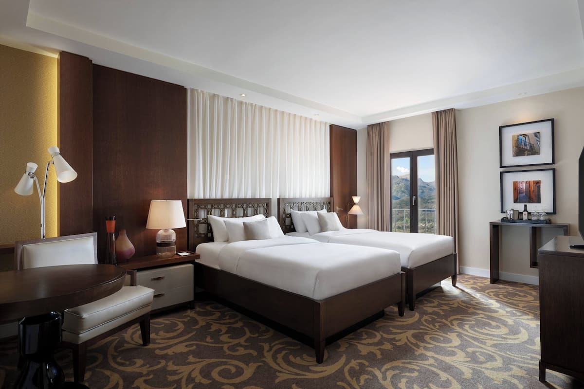A hotel room with two beds, a desk, a chair, a lamp, and a view of the outdoors.