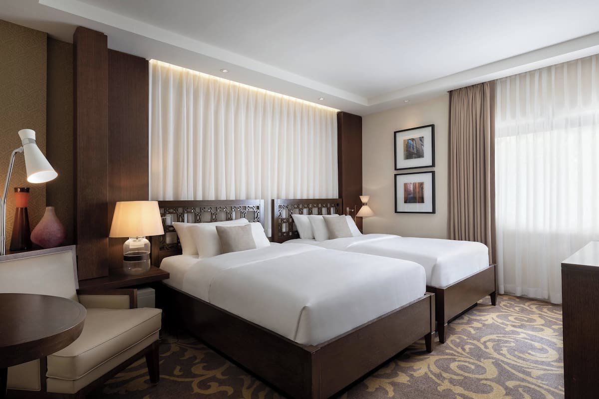 A hotel room with two beds, a desk, a chair, and a lamp.