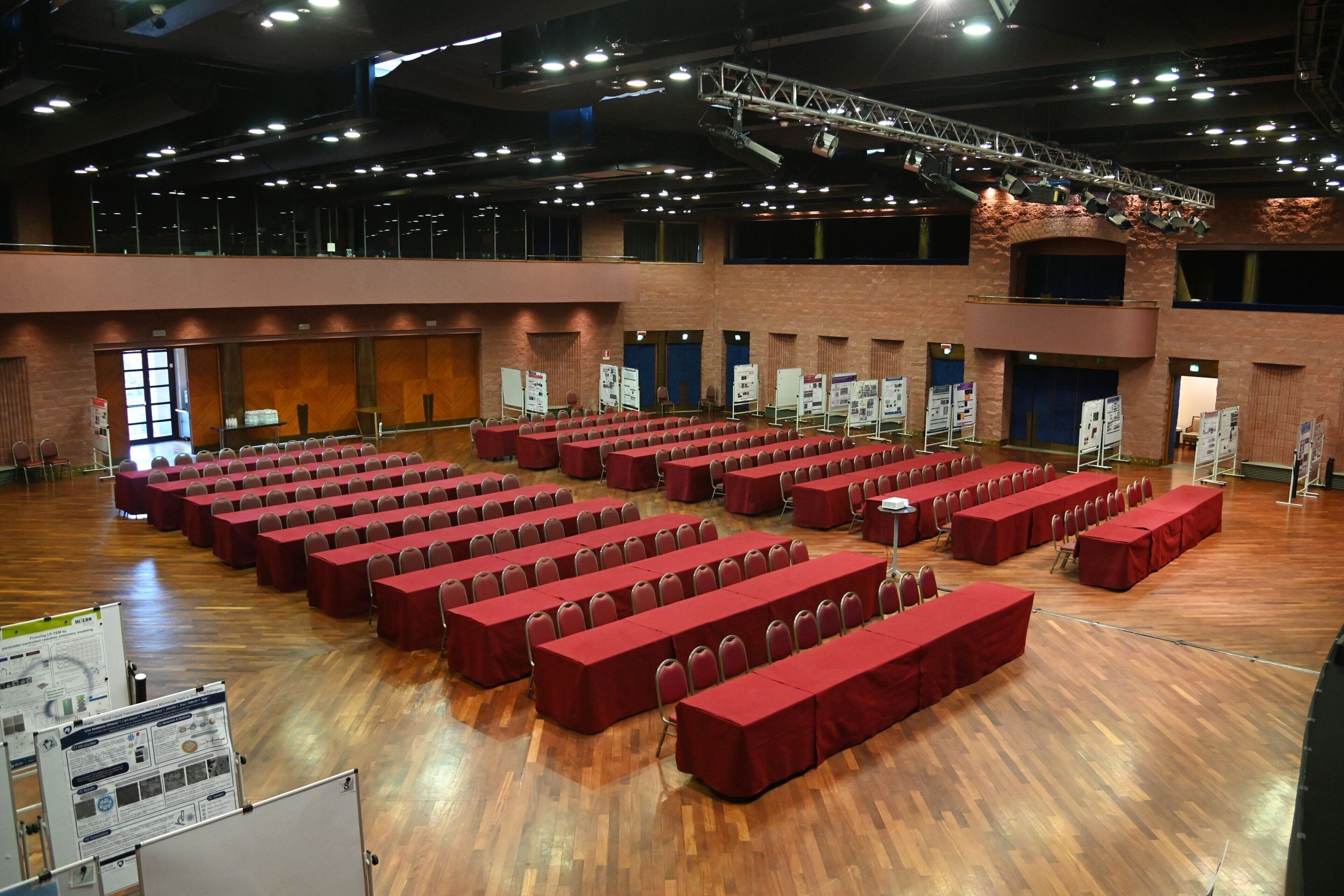 Une grande salle dotée de plusieurs rangées de tables et de chaises recouvertes de tissu rouge, d’un plancher en bois et d’un plafond élevé agrémenté de luminaires.