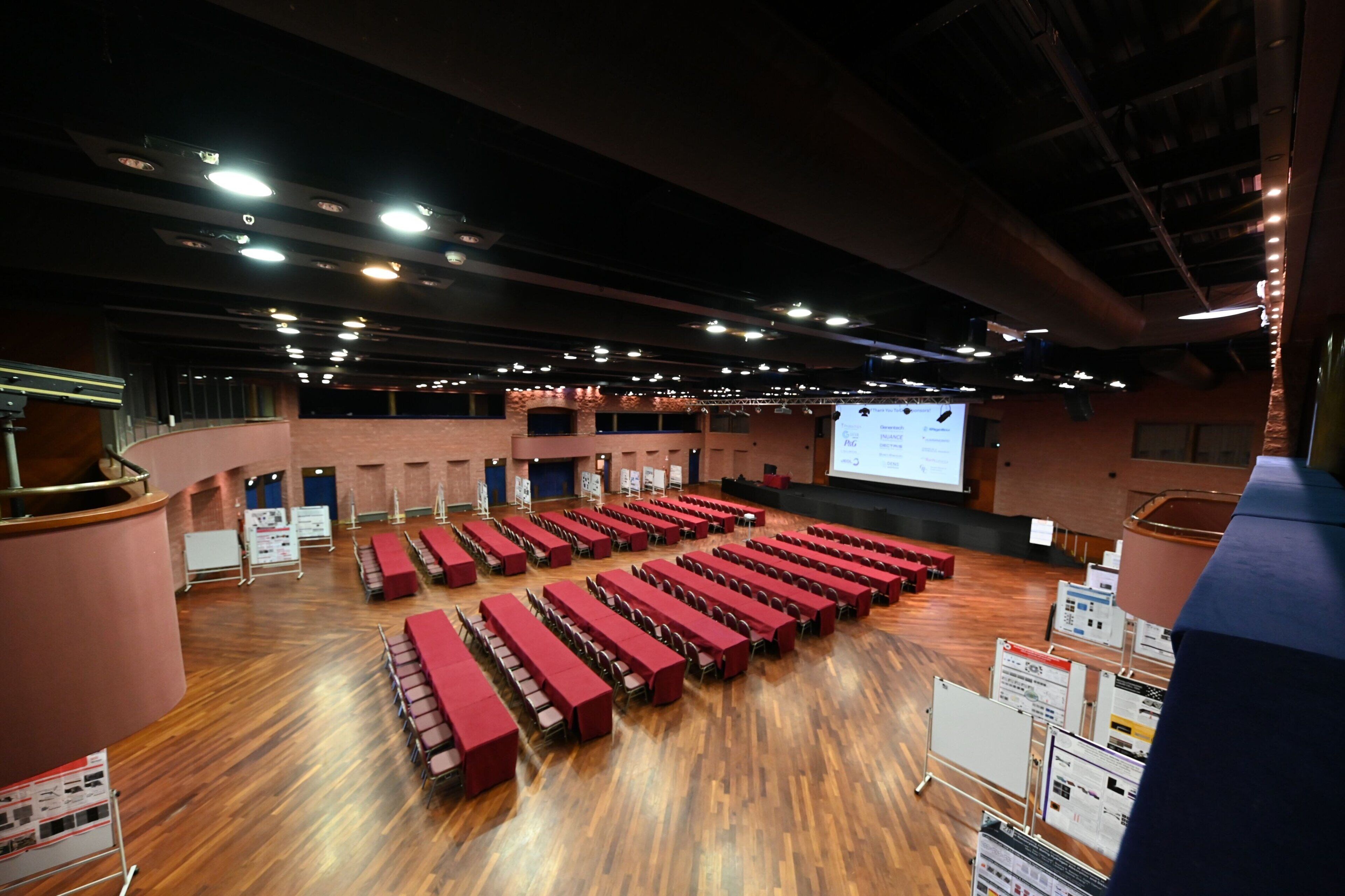 A large indoor space with rows of red tables and chairs, a stage with a screen, and a wooden floor.