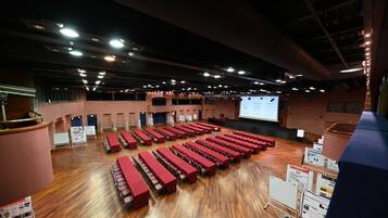 A large indoor space with rows of red tables and chairs, a stage with a screen, and a wooden floor.