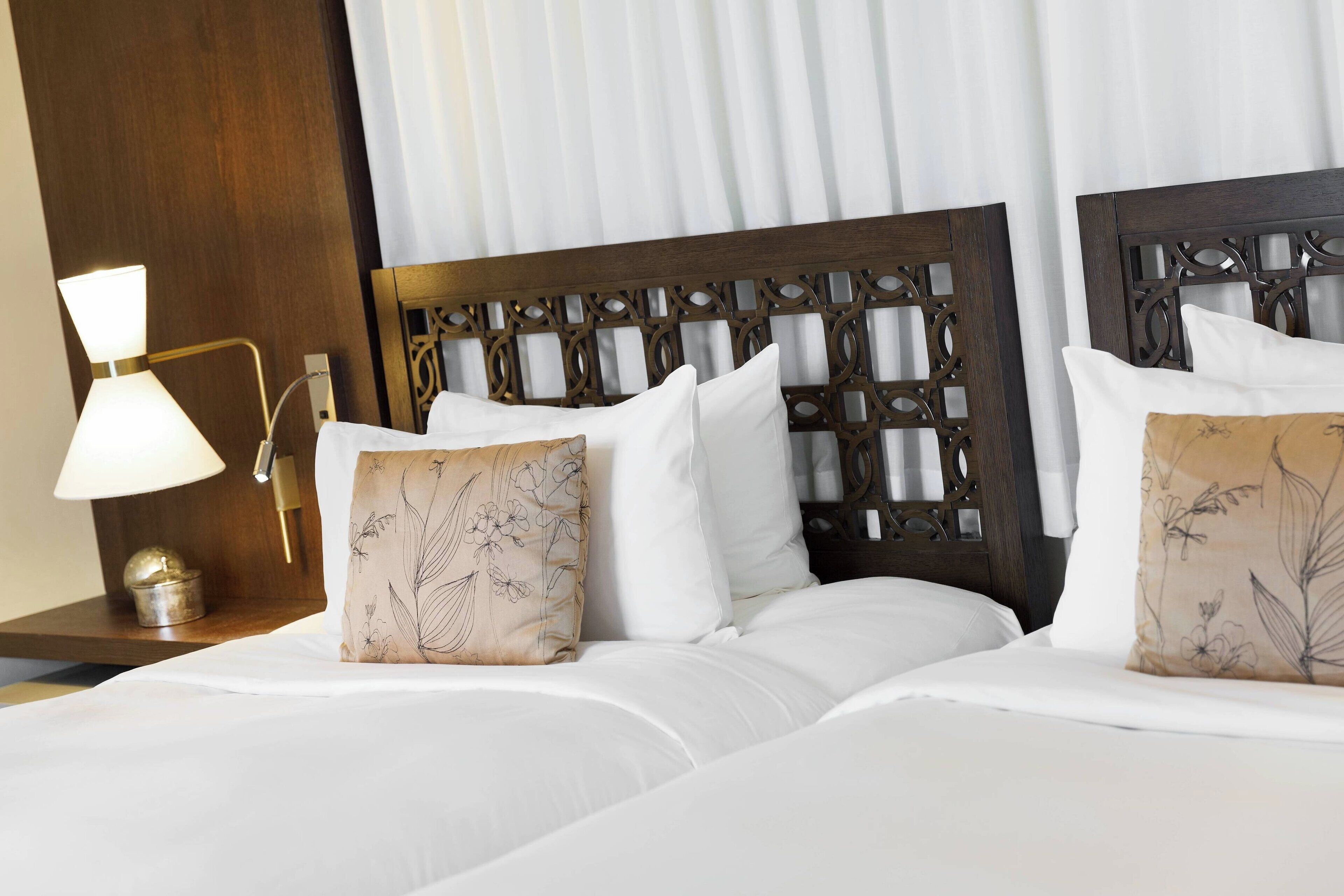 Een hotelkamer met twee bedden, een houten hoofdbord, een nachtkastje met lamp en een decoratief kussen.