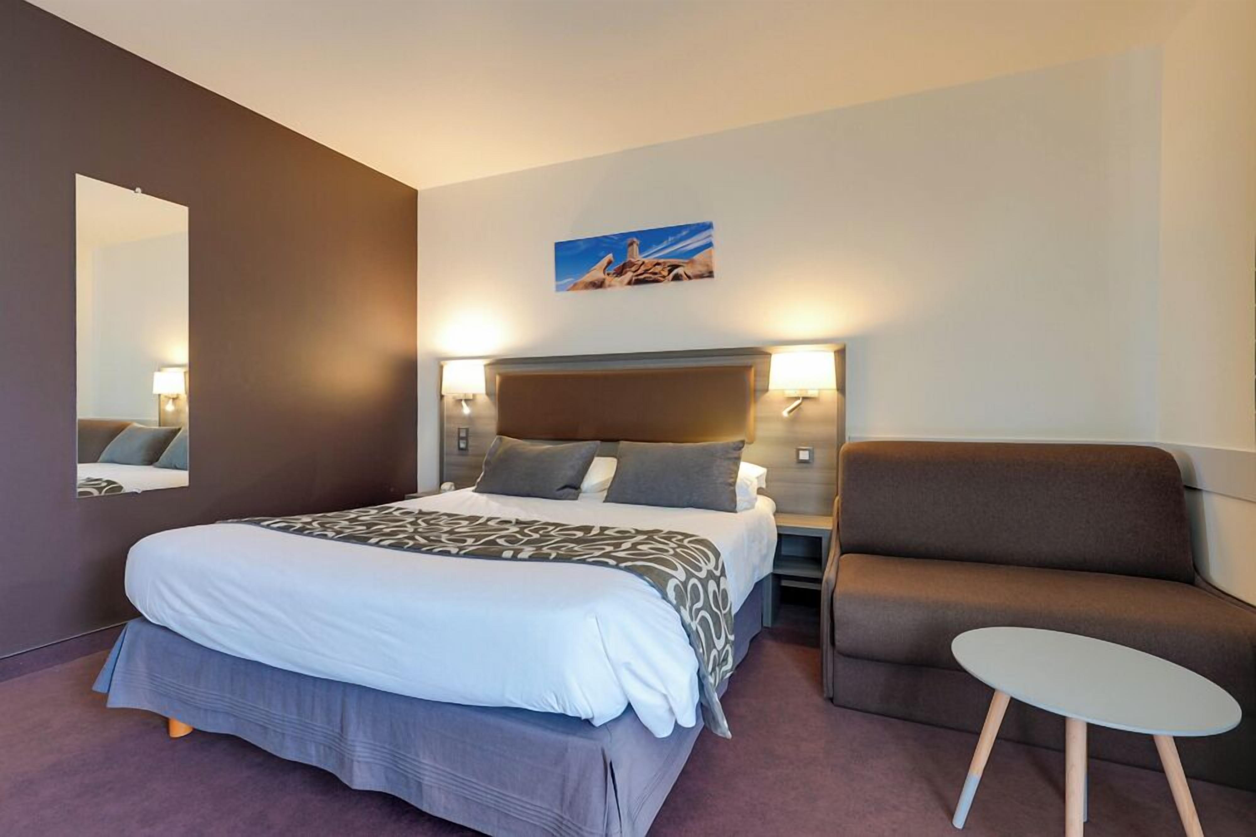 A hotel room with a large bed, a chair, a small table, a nightstand, a wall-mounted lamp, and a painting.