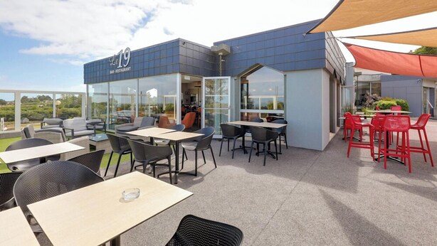 An outdoor seating area with tables and chairs, a glass facade, and a building with signage. - Golf de St Samson (Pleumeur-Bodou)