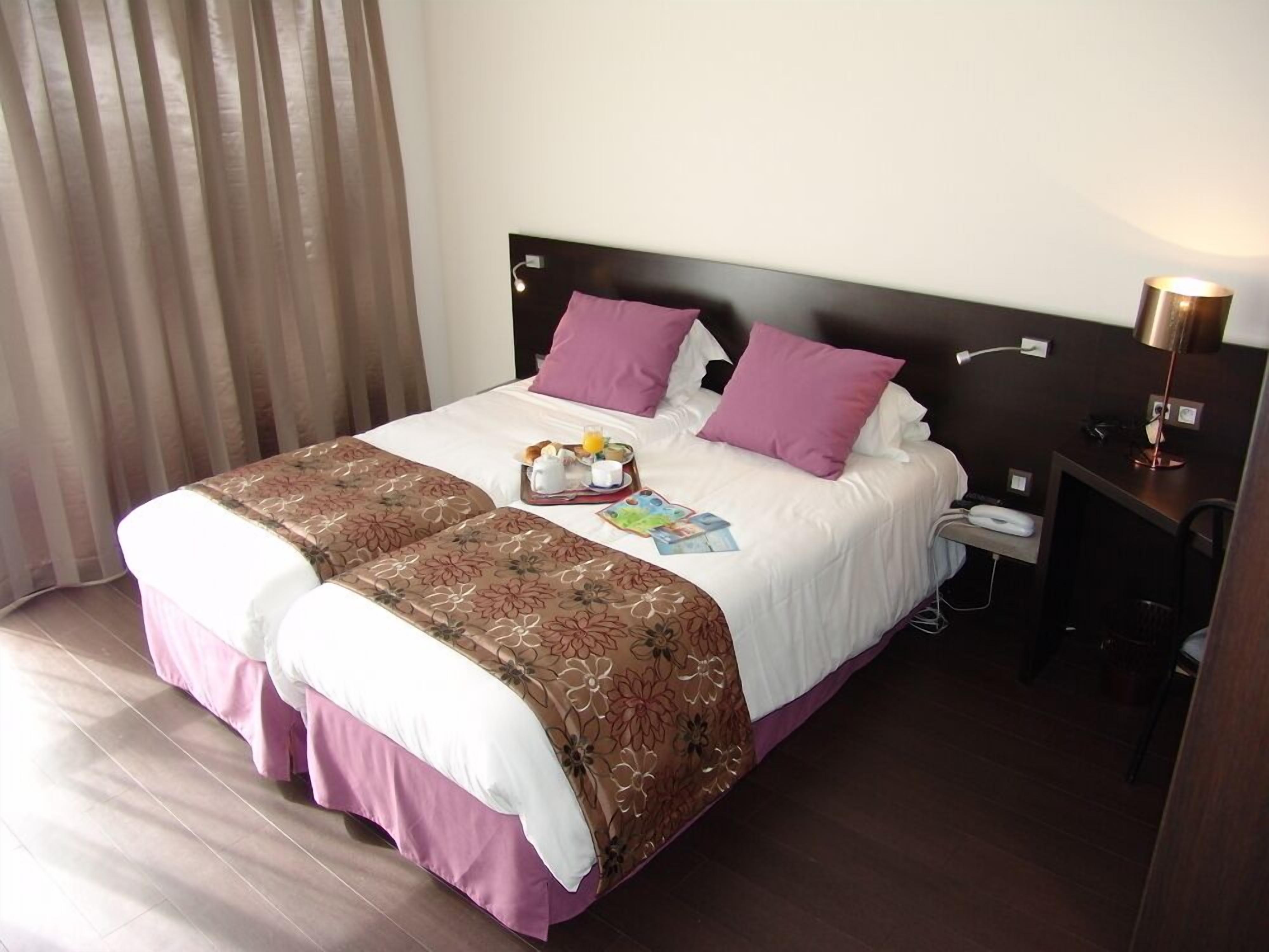 A hotel room with a bed, a bedside table with a lamp, and a window with curtains.