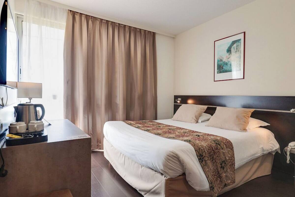Una habitación de hotel con una cama, una mesita de noche con lámpara, una cafetera, un cuadro en la pared y una ventana con cortinas.