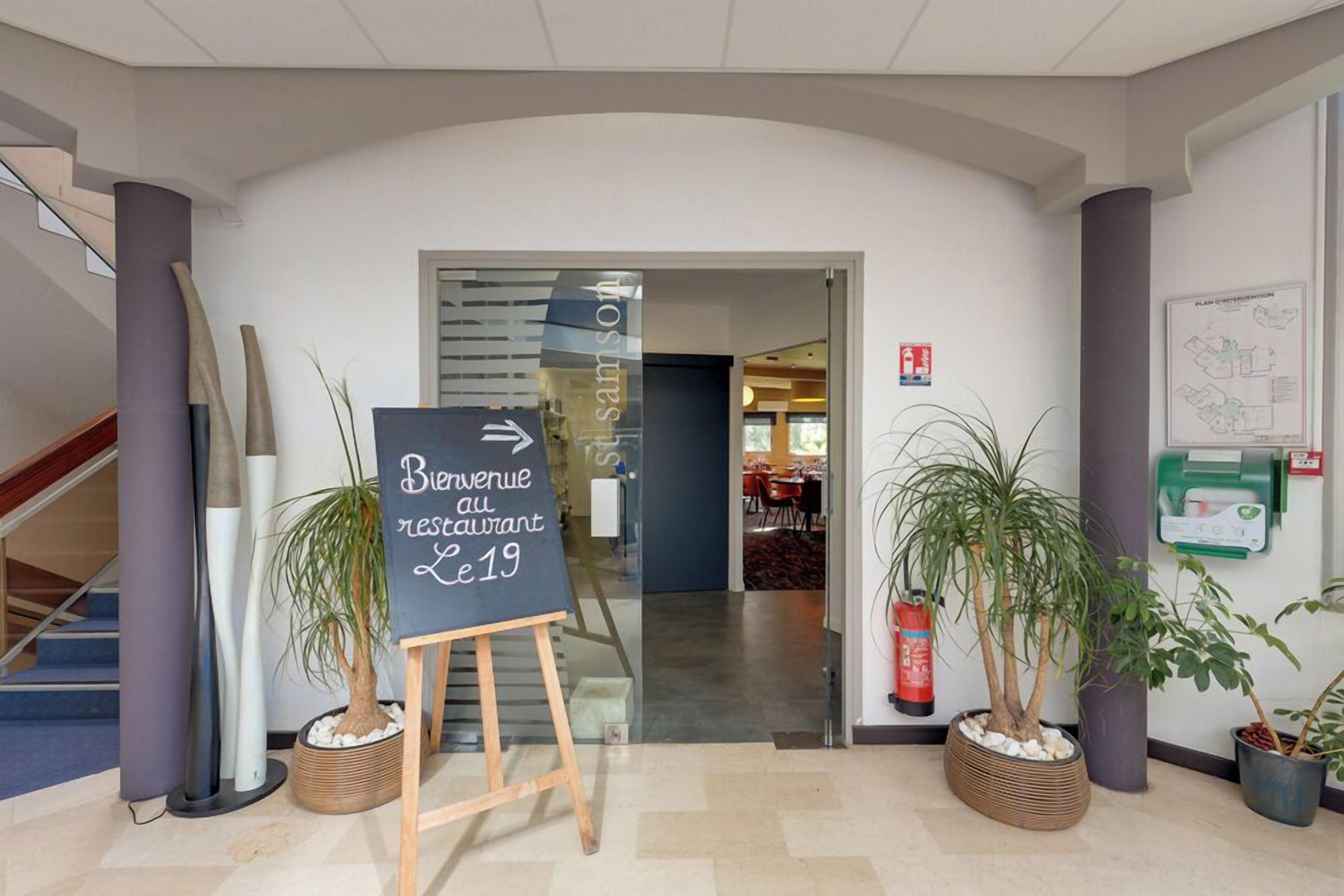 Un espace d’accueil avec une enseigne indiquant « Bienvenue au restaurant Le 19 », un escalier et des plantes en pot.