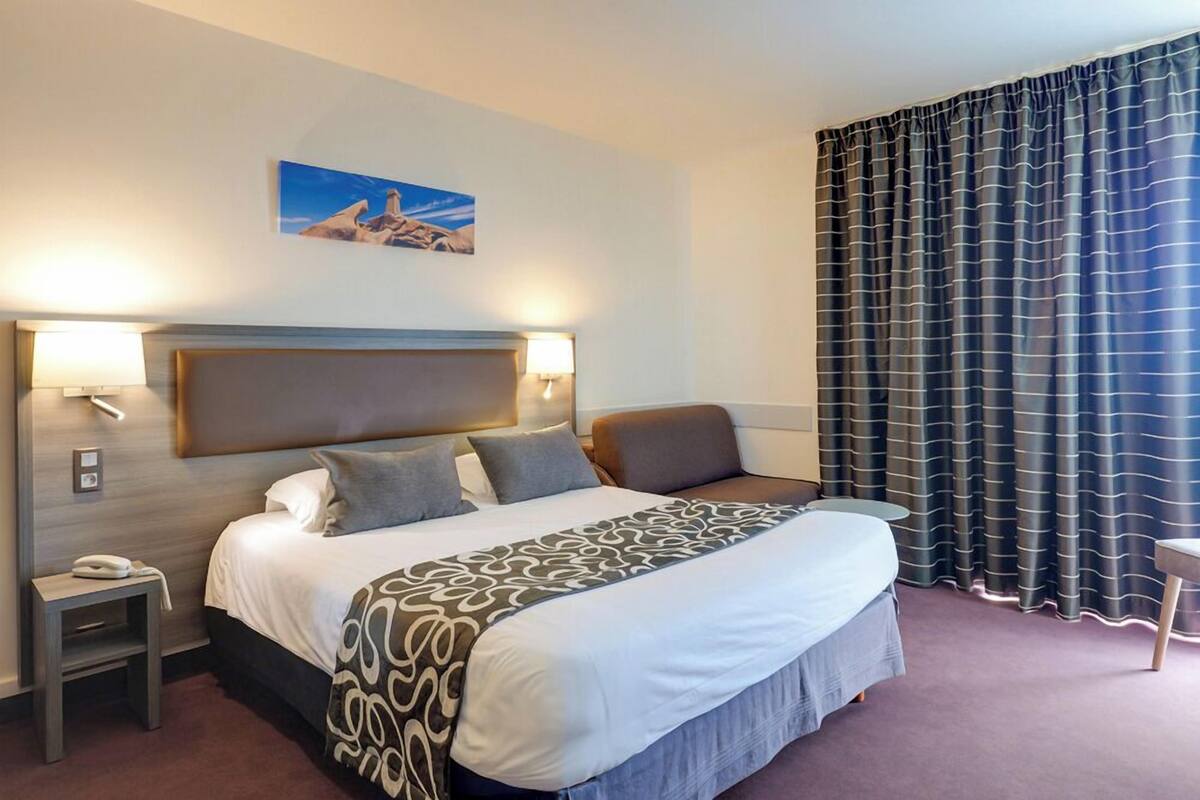 Habitación de hotel con una cama grande, lámparas de noche, una mesita, un teléfono, un cuadro en la pared y vistas al exterior a través de cortinas.