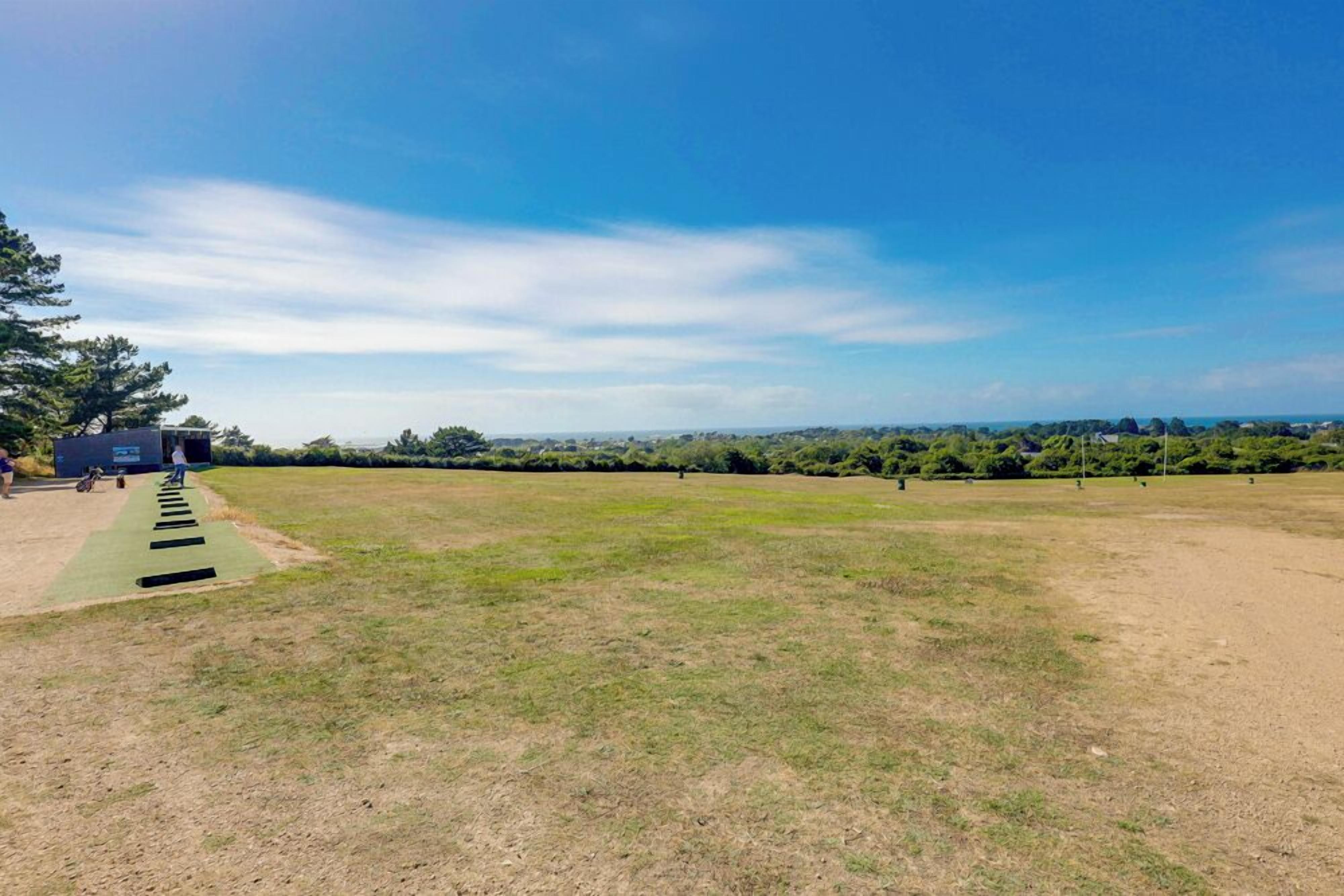 Un campo abierto y extenso con cielo despejado, un camino que lleva a un edificio y árboles a la distancia.