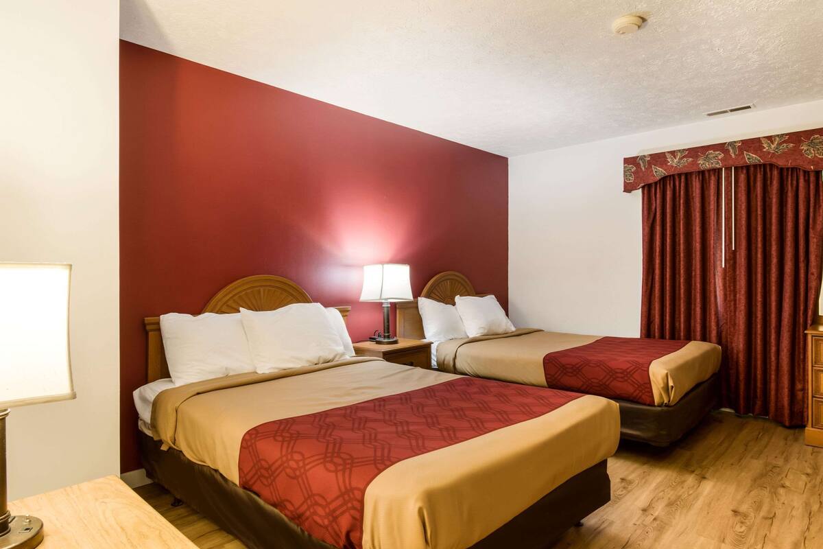 A hotel room with two beds, a wooden headboard, a nightstand with a lamp, and a red curtain.