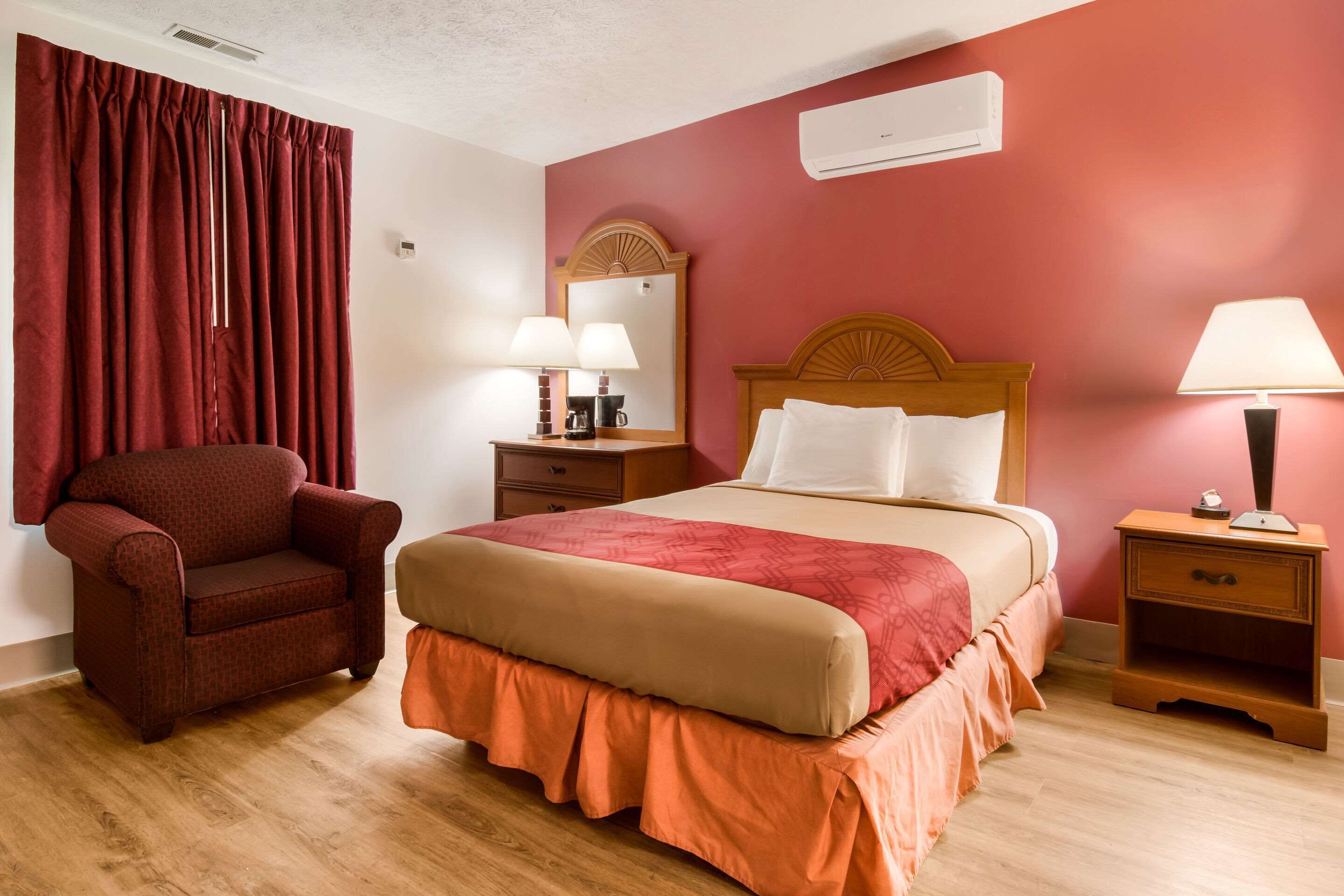 A hotel room with a bed, a chair, two bedside tables with lamps, a mirror, and an air conditioning unit on the wall.