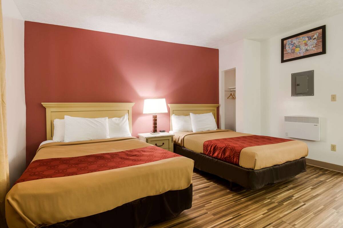 A hotel room with two beds, red walls, wooden flooring, a nightstand with a lamp, and a wall-mounted air conditioner.