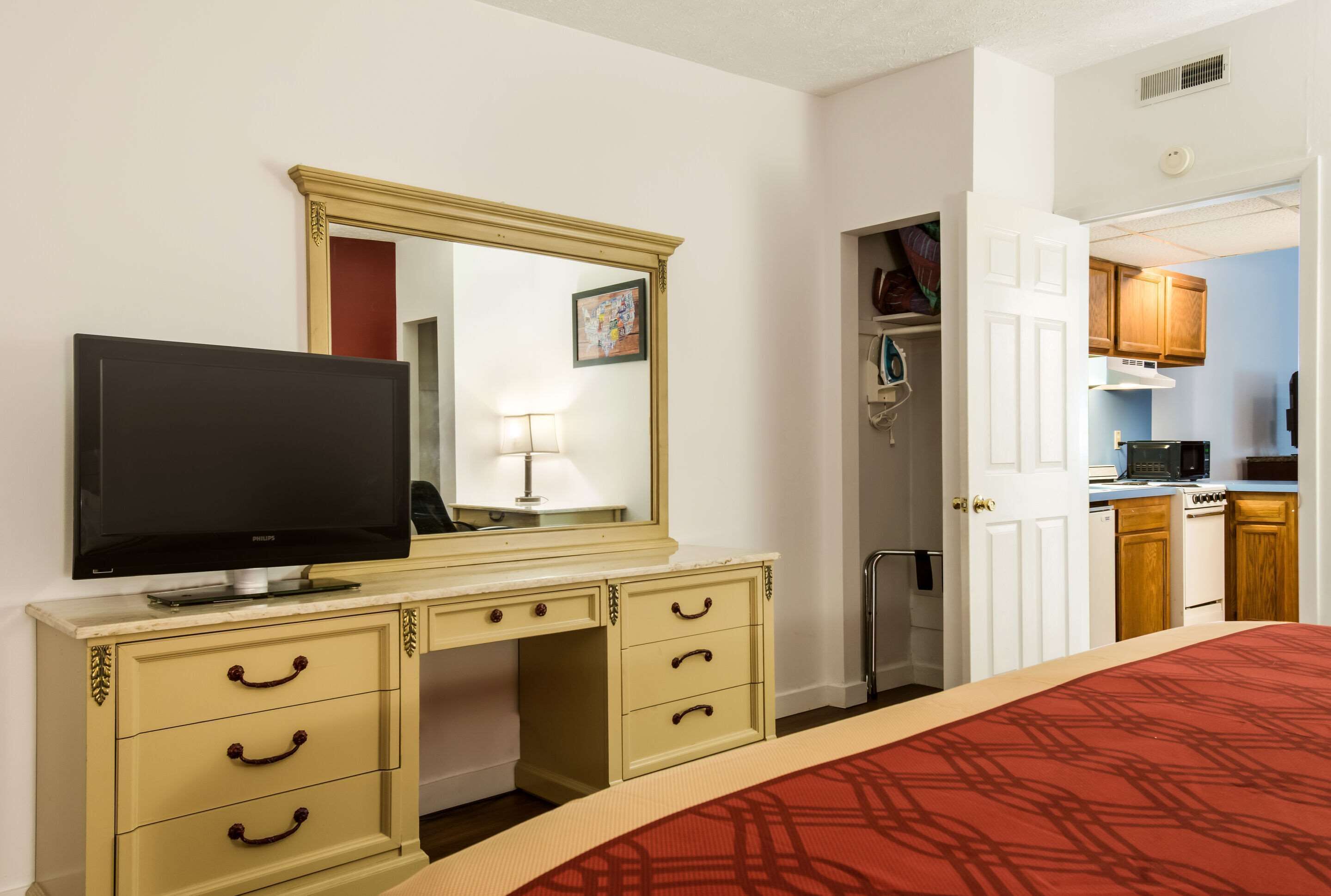 A hotel room with a bed, a television, a desk with drawers, a mirror, a lamp, and a kitchenette.