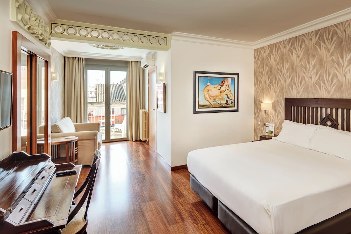 Ein Hotelzimmer mit einem großen Bett, einem Schreibtisch mit Klavier, einem Sessel und Blick auf die Stadt vom Balkon.