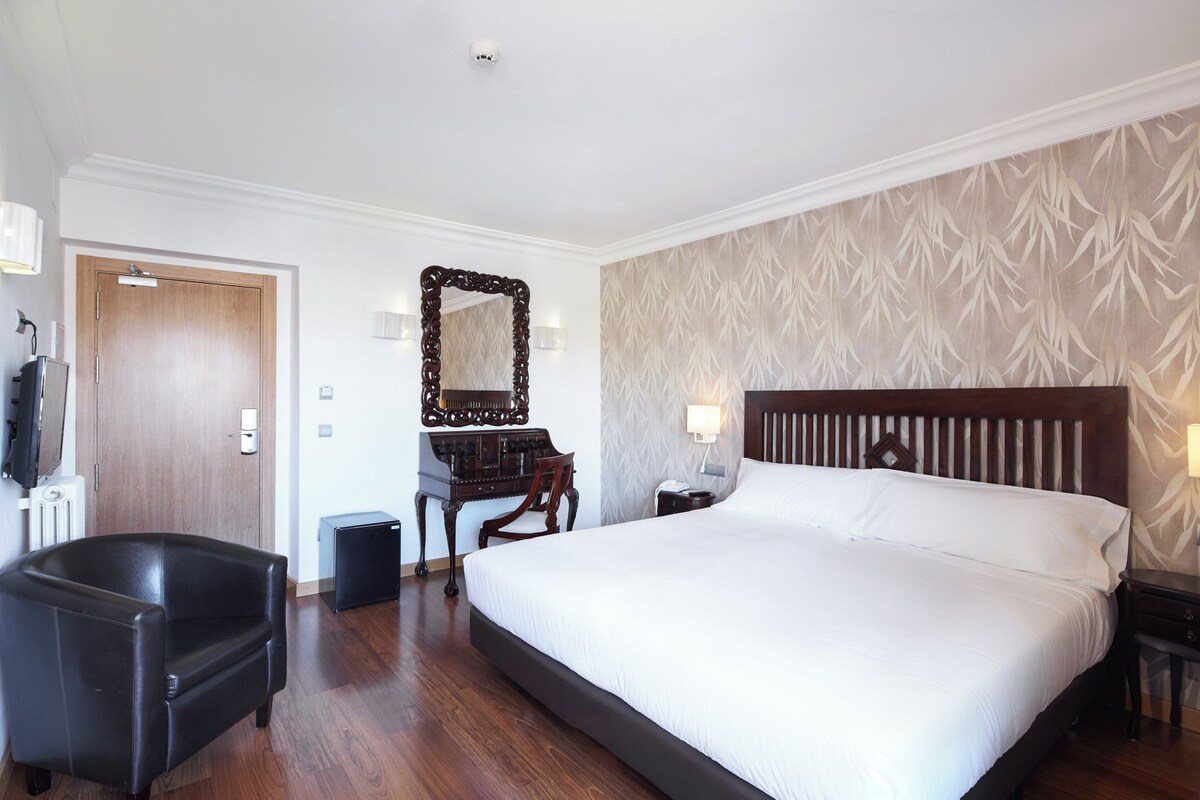A hotel room with a large bed, a wooden headboard, a desk with a chair, a mirror, and a radiator.