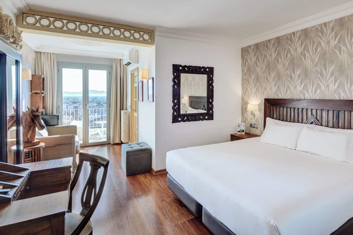 Ein Hotelzimmer mit einem großen Bett, einem Schreibtisch mit Stuhl, einem kleinen Hocker, einem Nachttisch mit Lampe und einem Spiegel an der Wand.