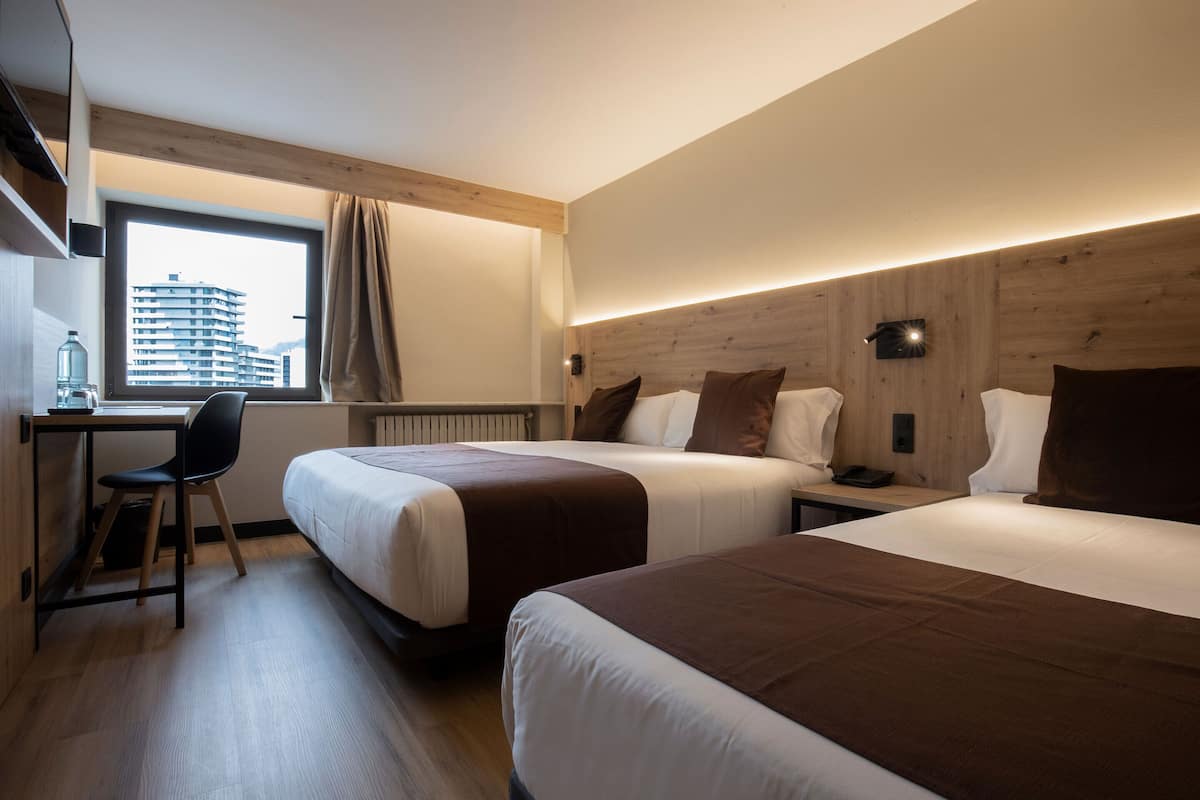 Hotellihuone, jossa on kaksi sänkyä, työpöytä, tuoli ja ikkunasta avautuva näkymä rakennukseen.