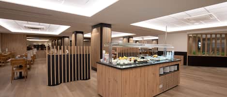 A modern hotel lobby with a central breakfast bar, wooden flooring, and a seating area with tables and chairs.