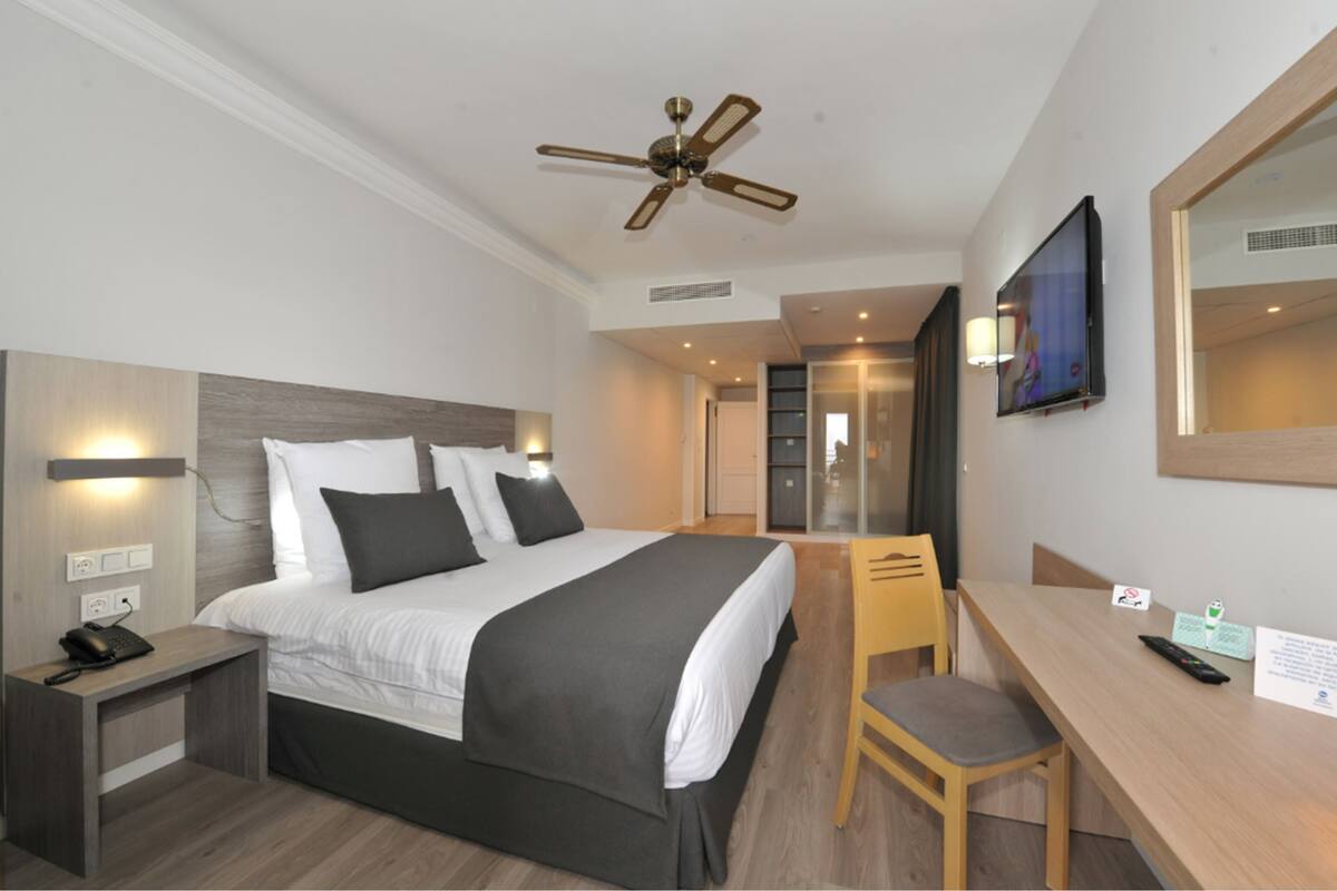 A hotel room with a bed, bedside table, chair, desk, TV, and ceiling fan.