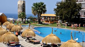 Zona de piscina con sombrillas de paja, sillas de descanso y vista al mar.