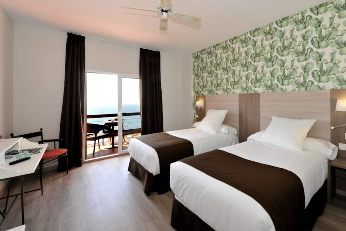 A hotel room with two beds, a desk, and a view of the sea.