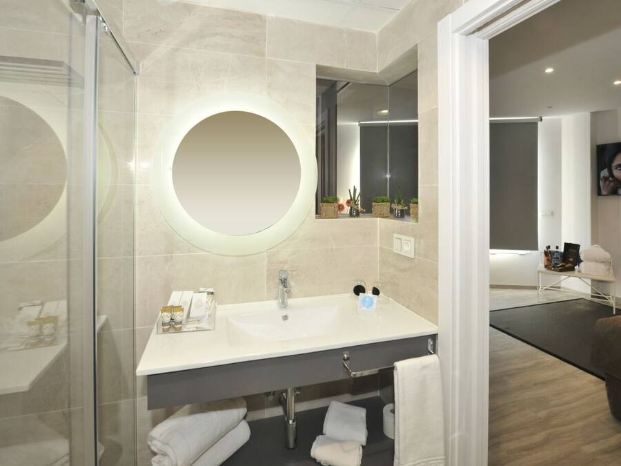 Une salle de bain moderne avec une douche à l’italienne en verre, un miroir rond, un lavabo blanc avec des articles de toilette et un coin salon aux couleurs sombres.