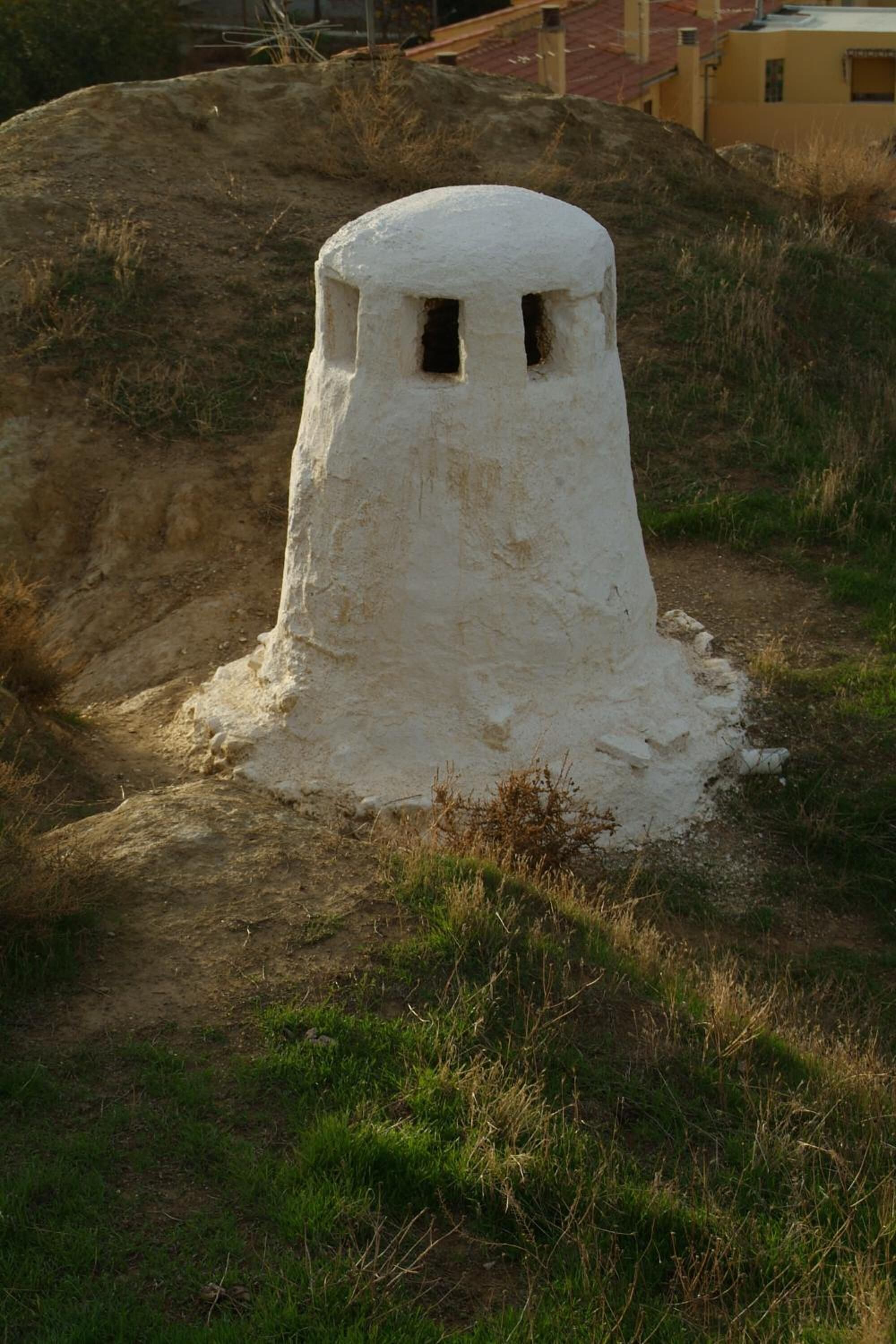 Una estructura blanca y cilíndrica con una pequeña abertura en la parte superior, ubicada en una colina cubierta de hierba con un sendero de tierra que lleva hacia ella.