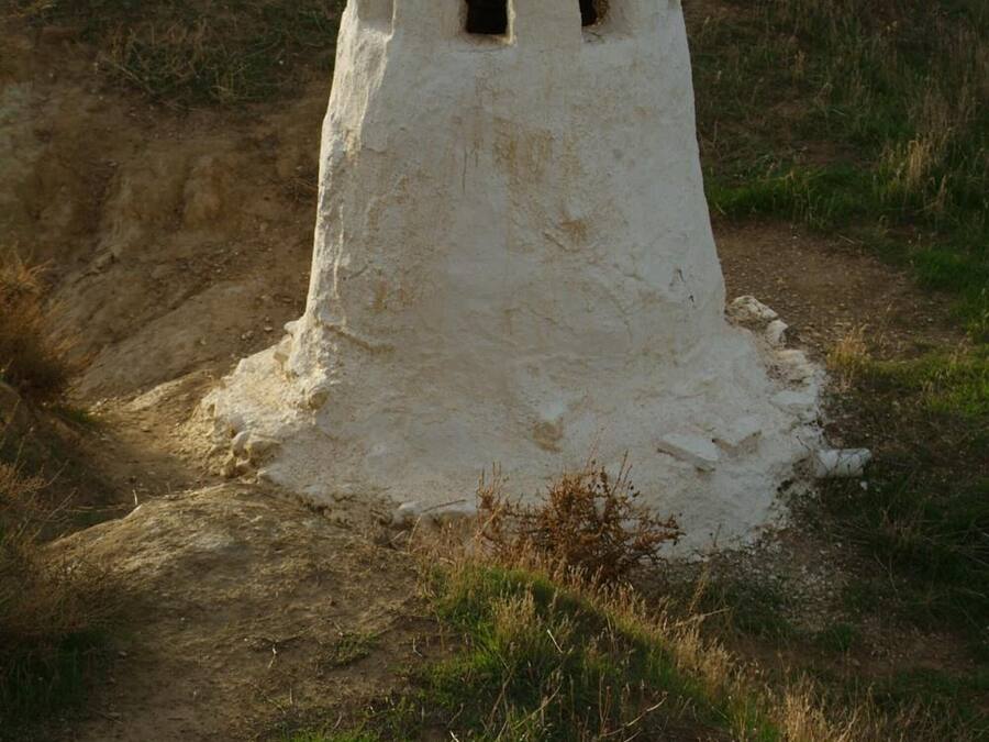 A white, cylindrical structure with a small opening at the top, situated on a grassy hill with a dirt path leading up to it.
