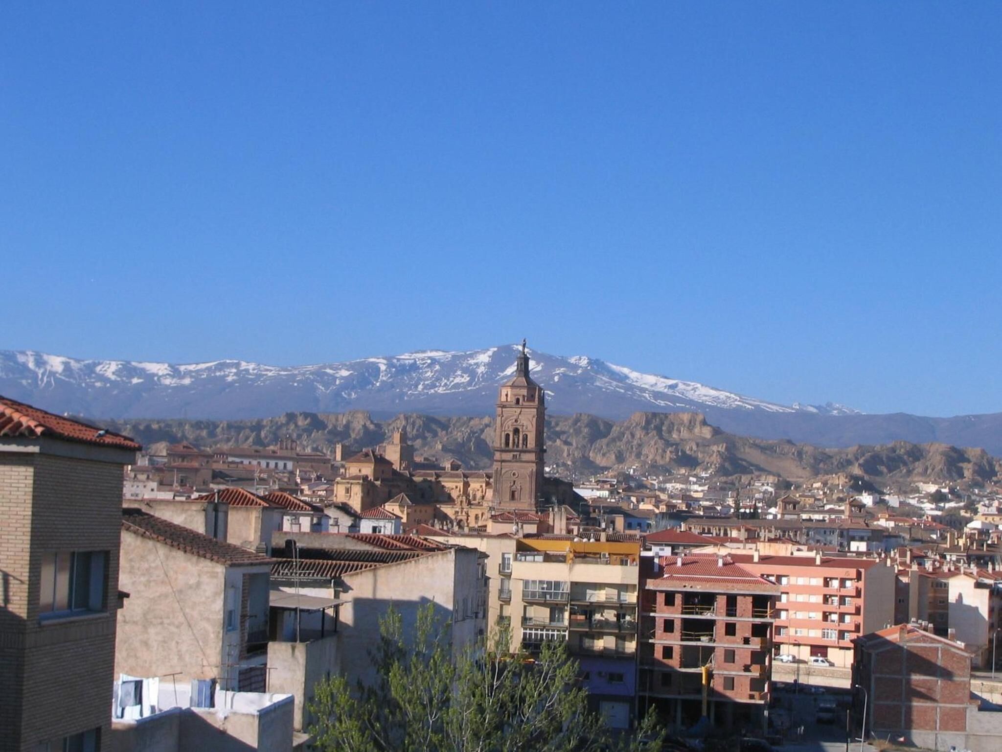 Un paisaje urbano con una torre de iglesia destacada, edificios residenciales y una montaña nevada al fondo.