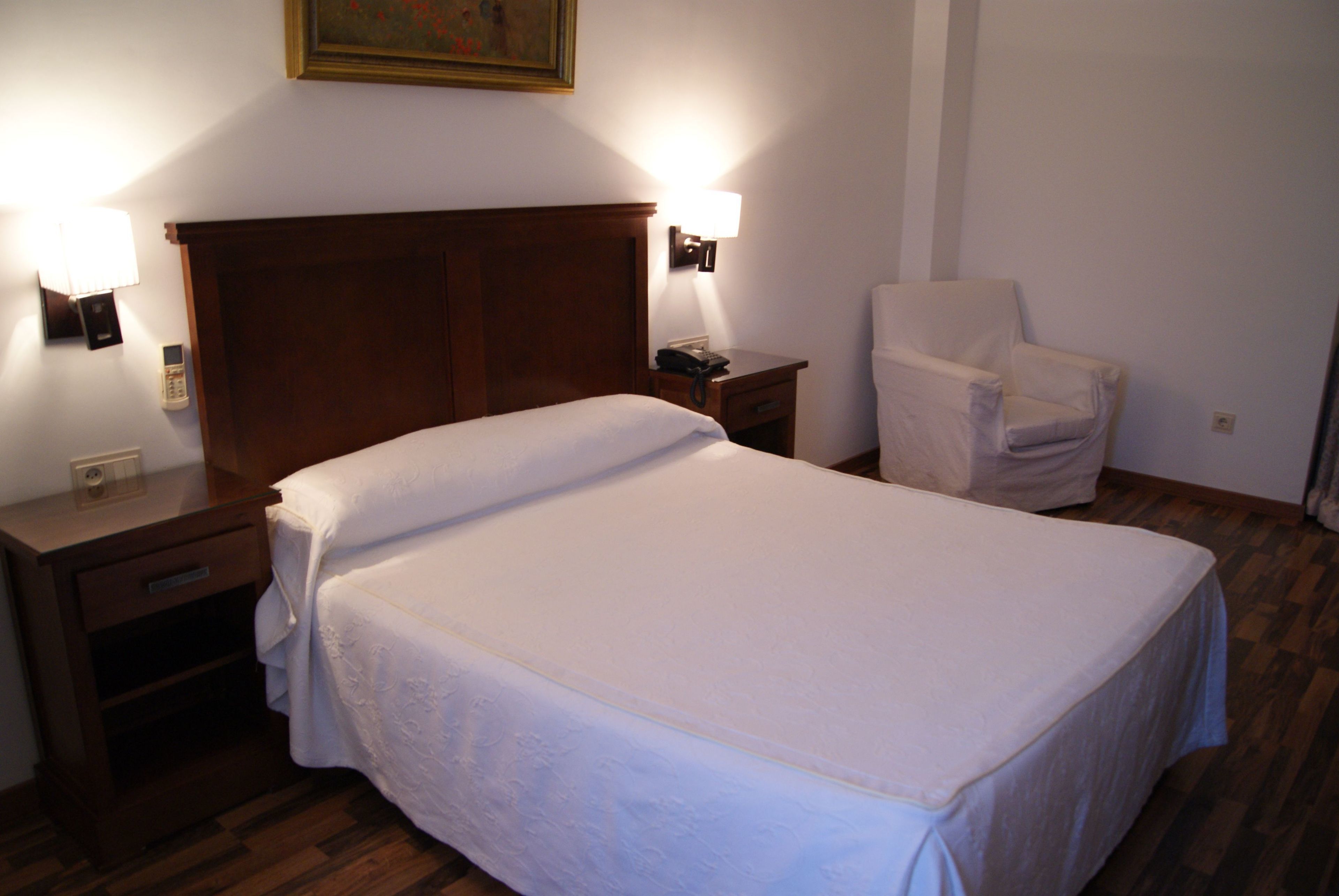Una cama bien hecha con cabecera de madera, una mesita de noche con lámpara y una silla en una habitación de hotel.