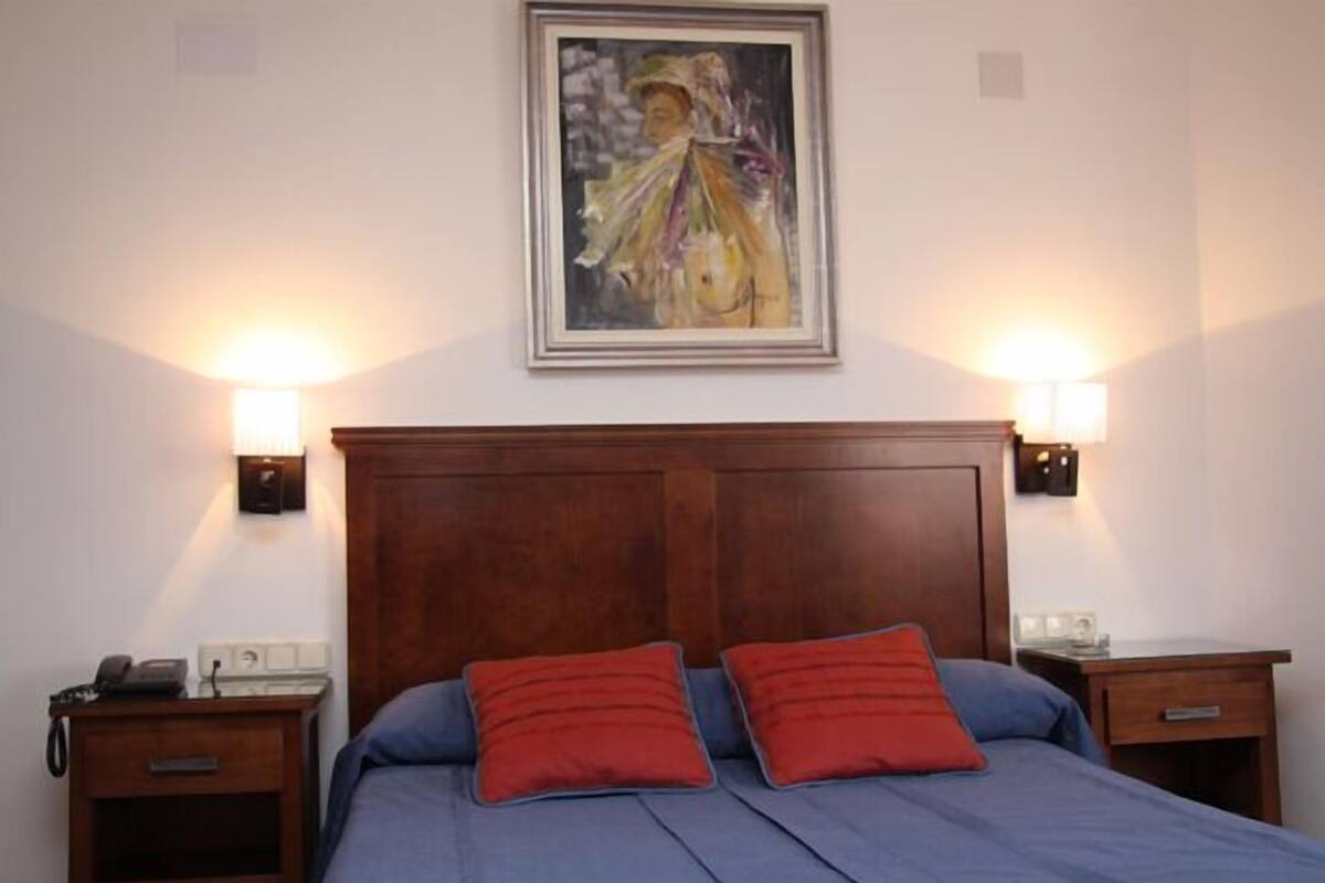 Ein Schlafzimmer mit einem Holzkopfteil, zwei Nachttischen, einem Bett mit blauer Bettwäsche und roten Kissen sowie einem Bild an der Wand.