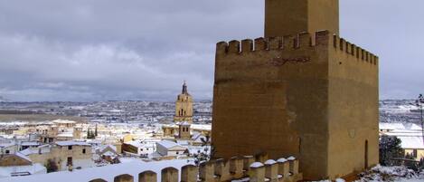 Una torre histórica con almenas, rodeada por un muro cubierto de nieve y un pueblo con edificios al fondo.