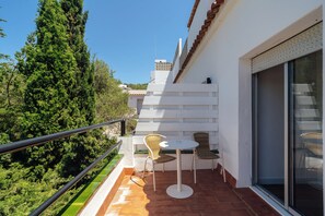 A balcony with two wicker chairs and a small round table, overlooking a lush green landscape and a clear blue sky.